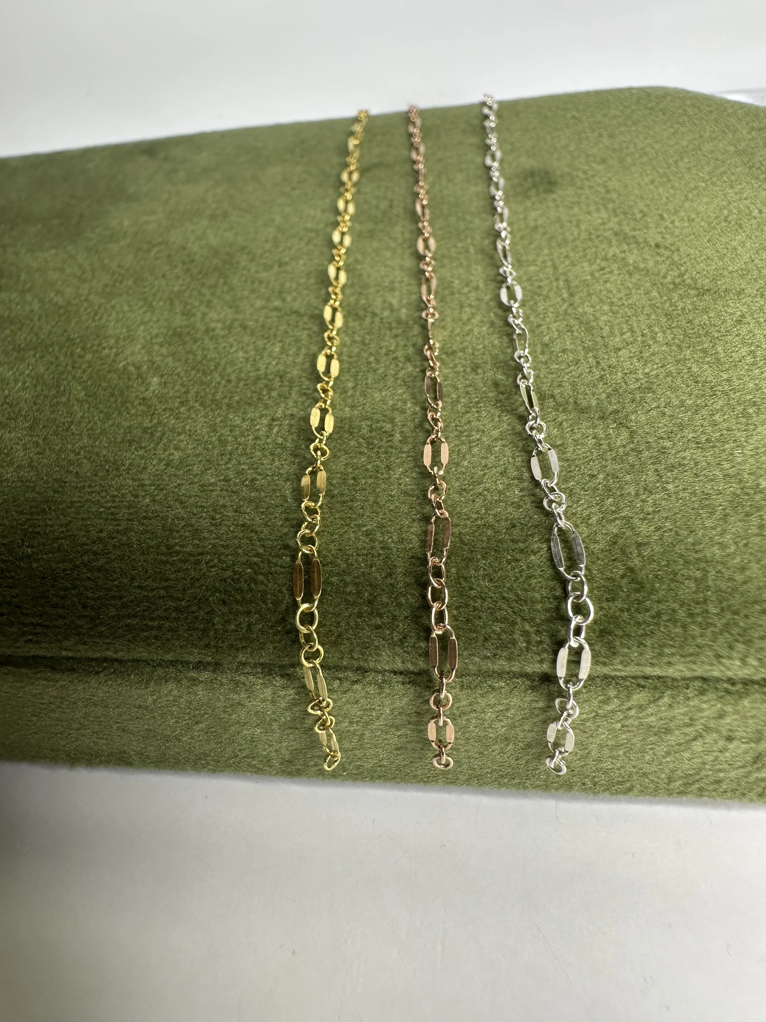 Three metal chain necklaces, one gold, one rose gold, and one silver, displayed on a green velvet cushion.