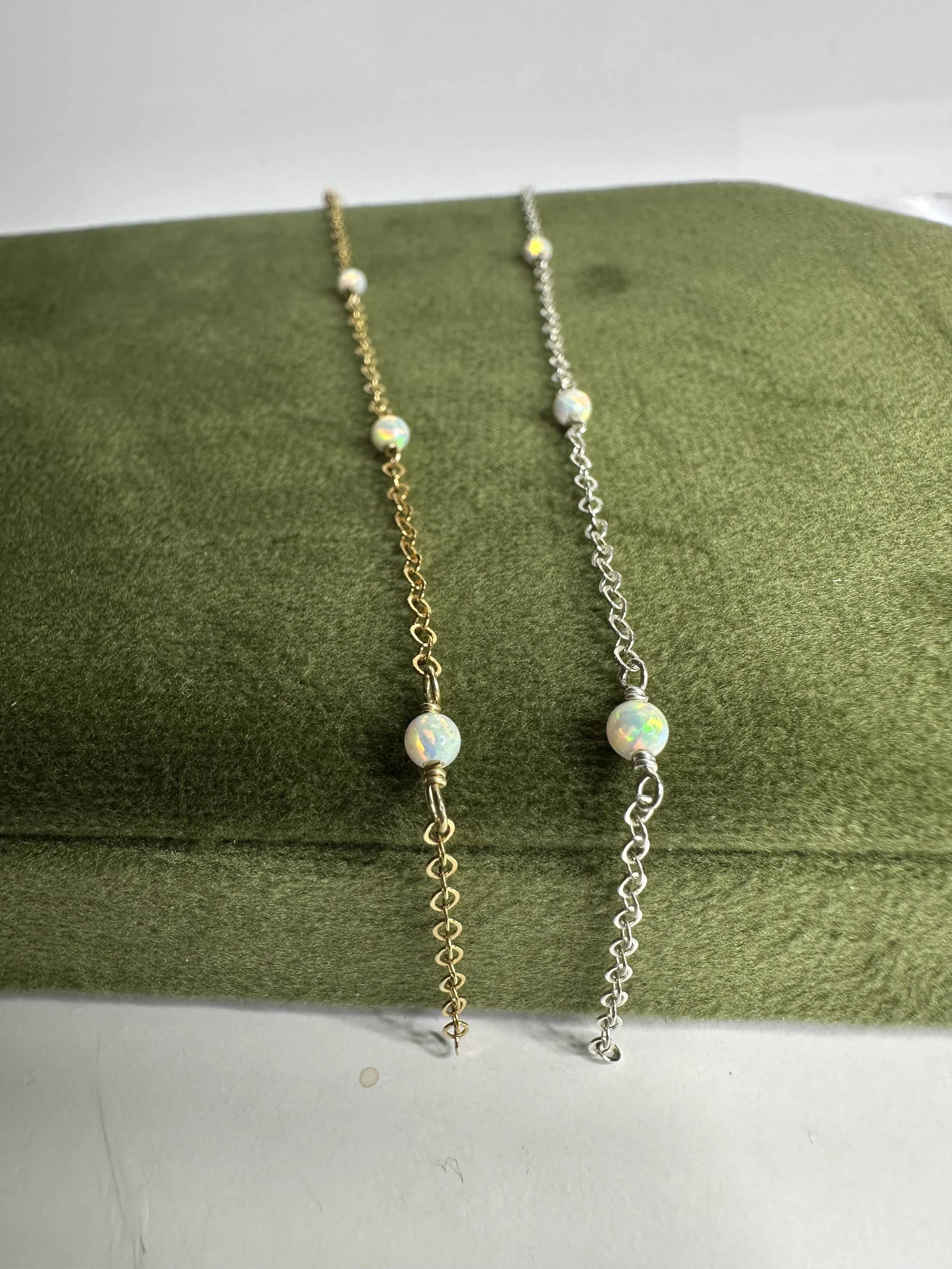 Two delicate necklaces with iridescent beads, one on a gold chain and the other on a silver chain, displayed on a green textured surface.