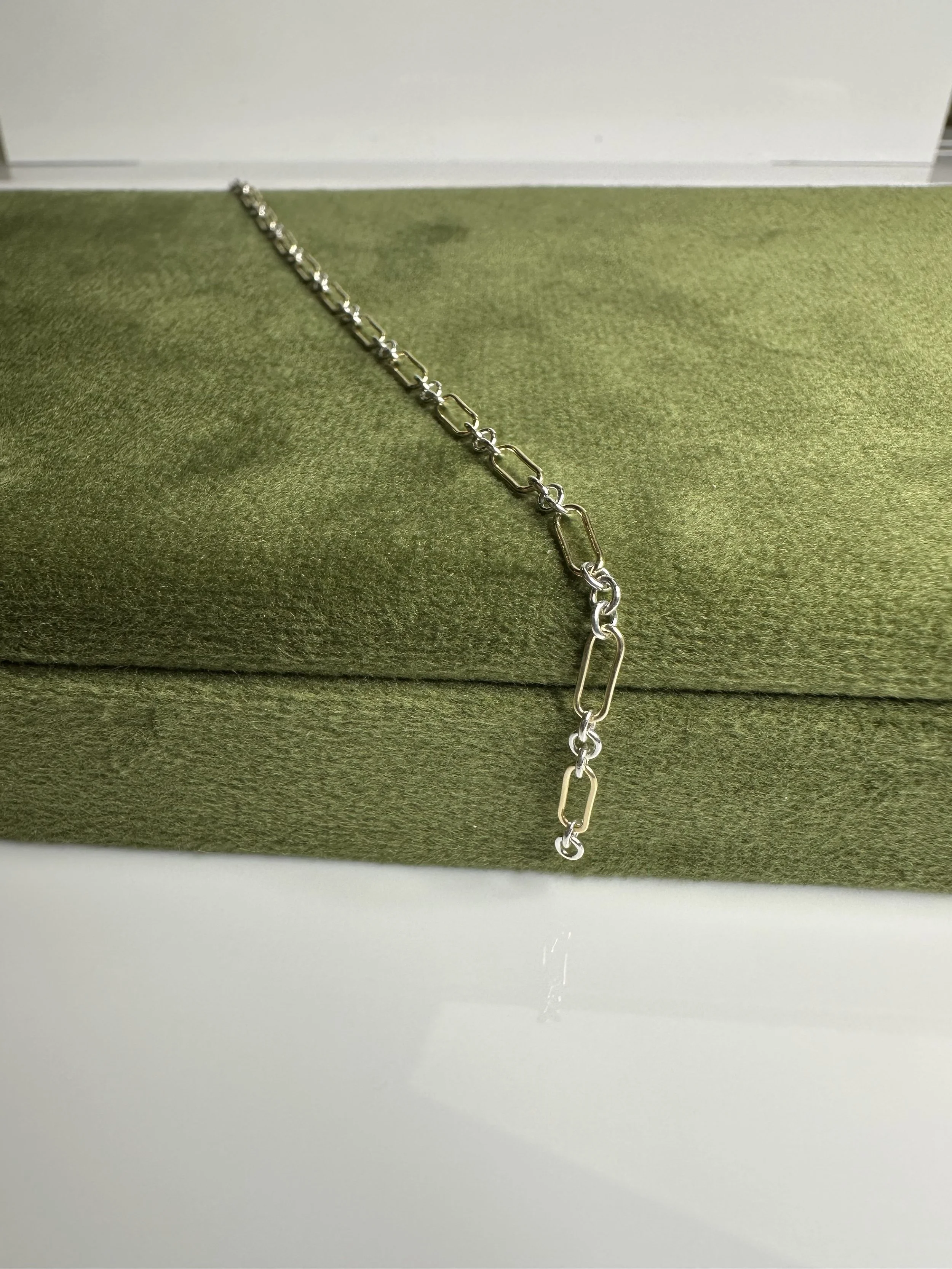 A silver and gold chain necklace displayed on a green velvet cushion.