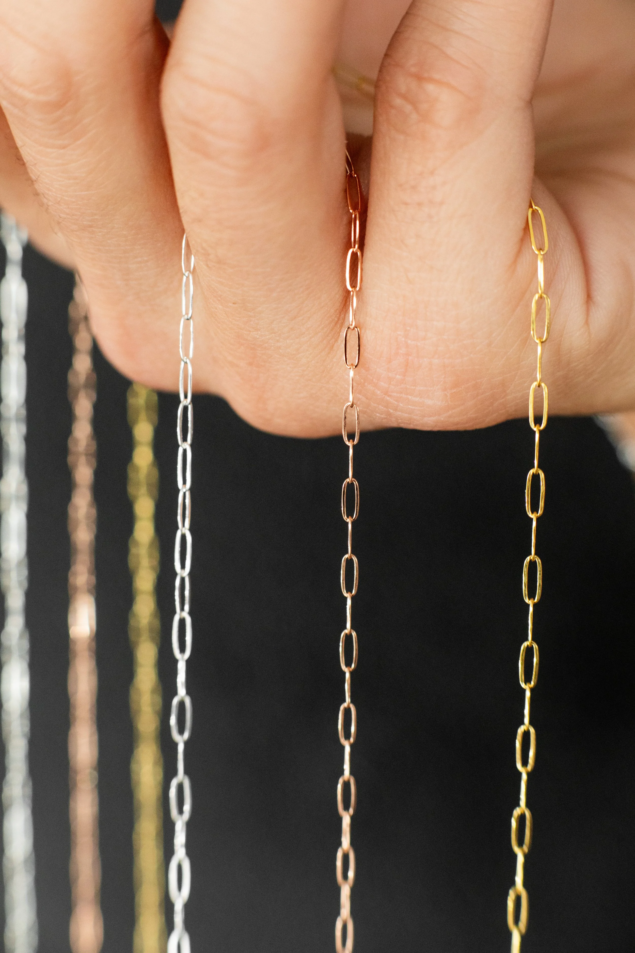 Hand holding three thin chain necklaces in silver, rose gold, and gold, against a black background.