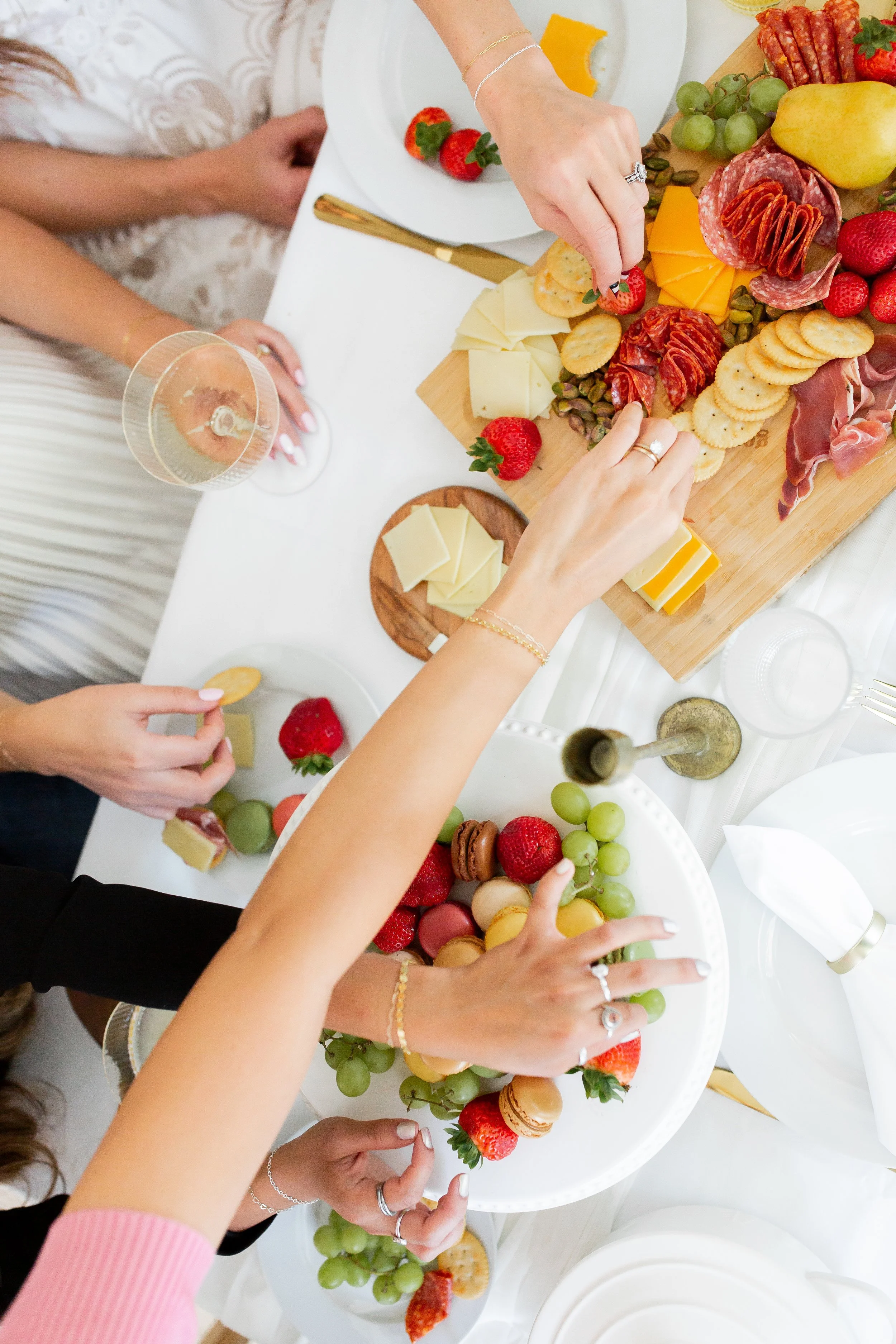 People gathering around a buffet table with cheese, crackers, strawberries, grapes, macarons, and charcuterie, reaching for food.