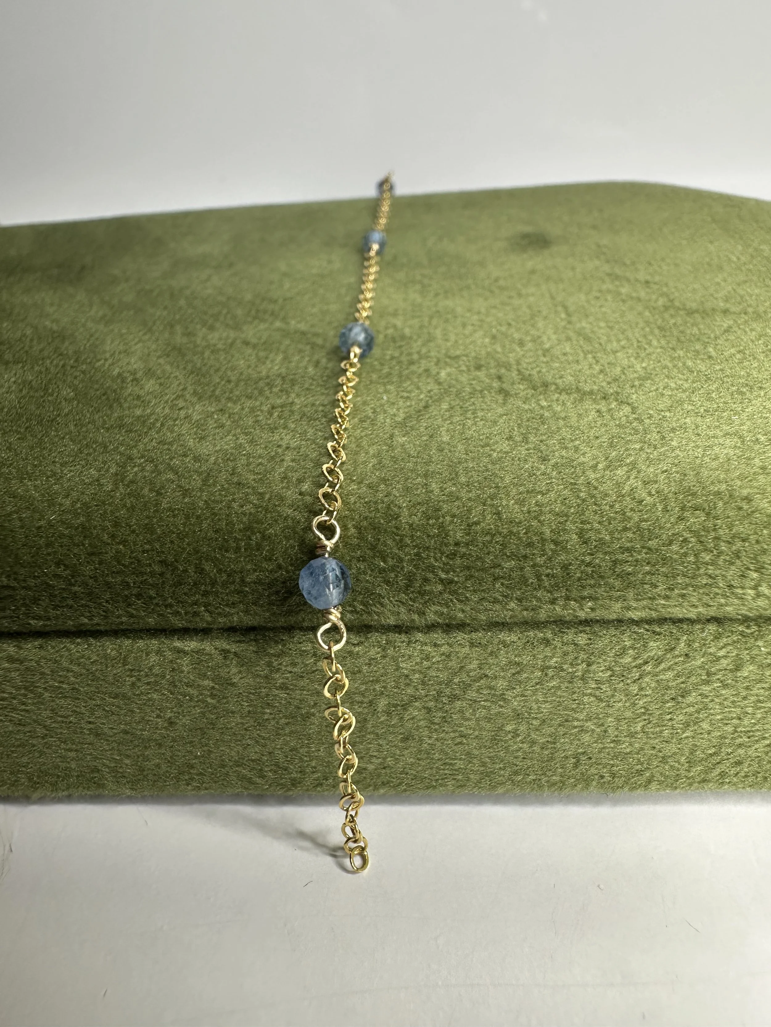 Gold chain bracelet with blue beads on a green fabric display cushion.