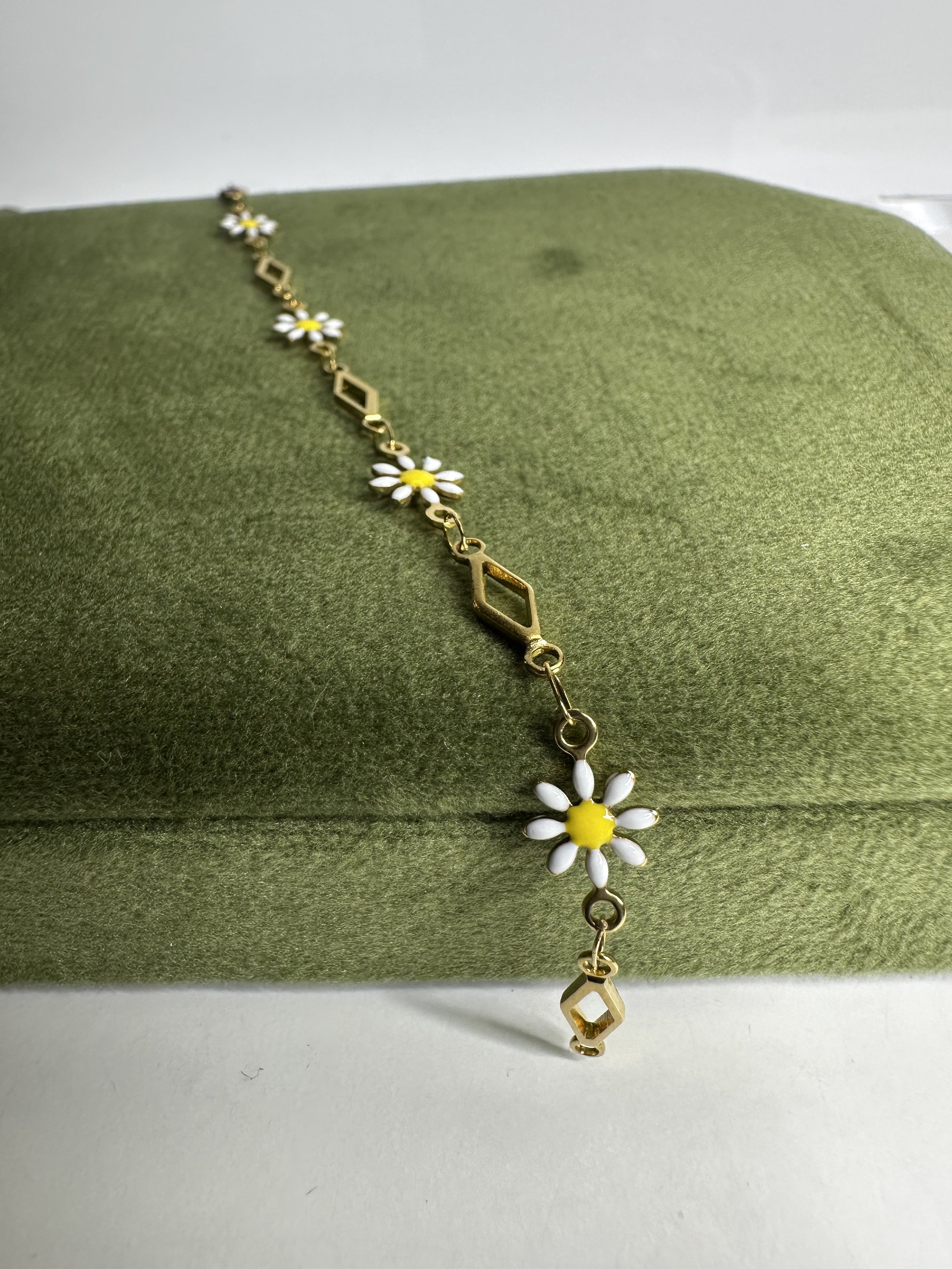 Gold necklace with white daisy flowers and geometric gold links, draped over a green textured fabric