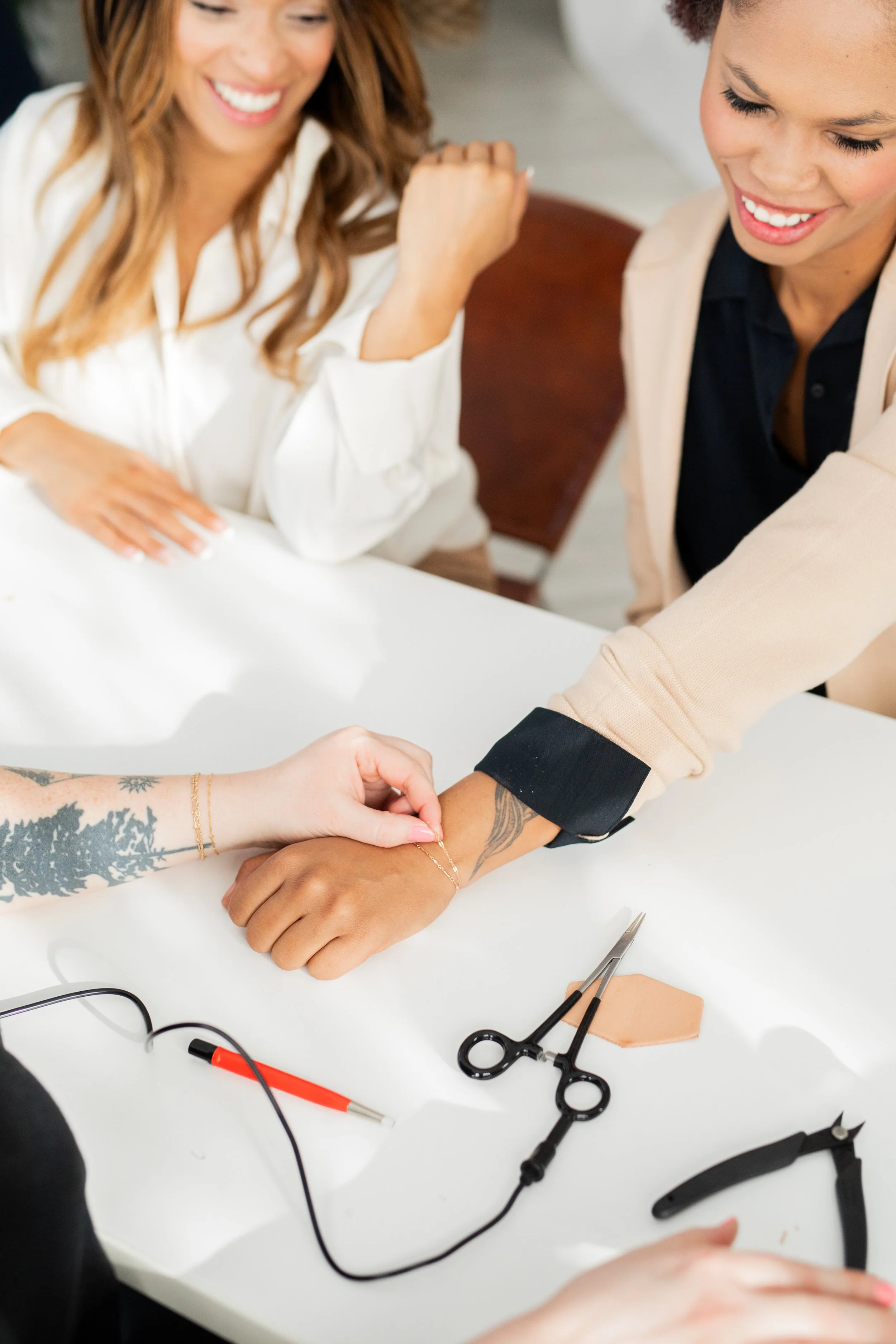 Two women observe as a third woman puts a necklace on her wrist at a jewelry-making workshop. Jewelry tools and equipment are on the table, and the women are smiling and appear happy.