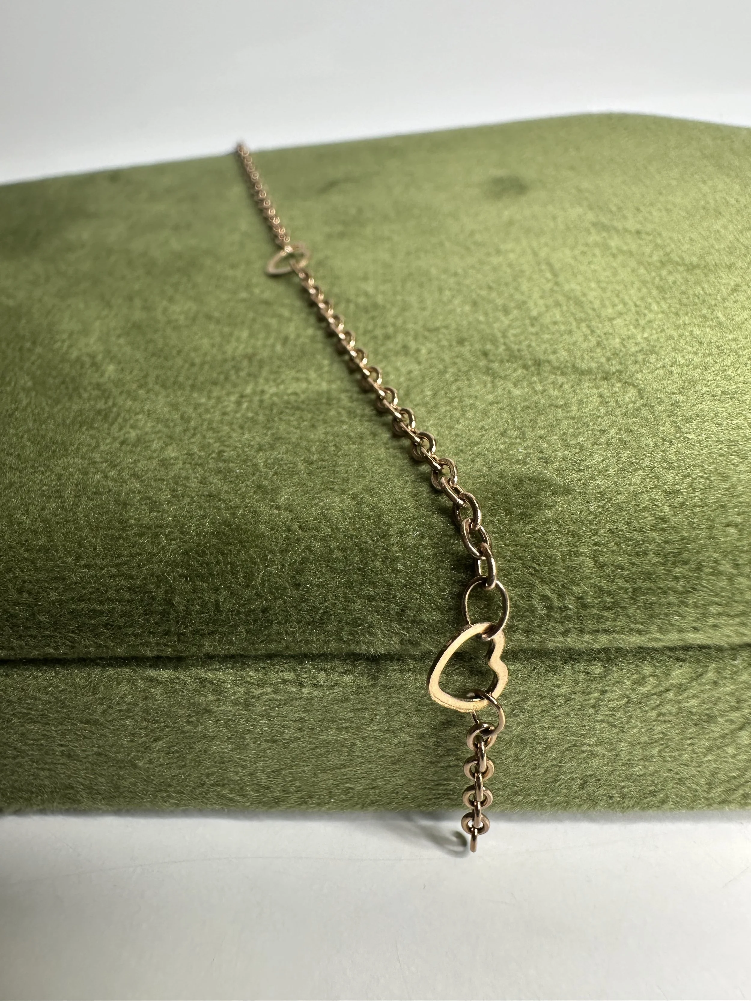 A gold chain bracelet with a heart-shaped charm, draped over a green, textured jewelry display.