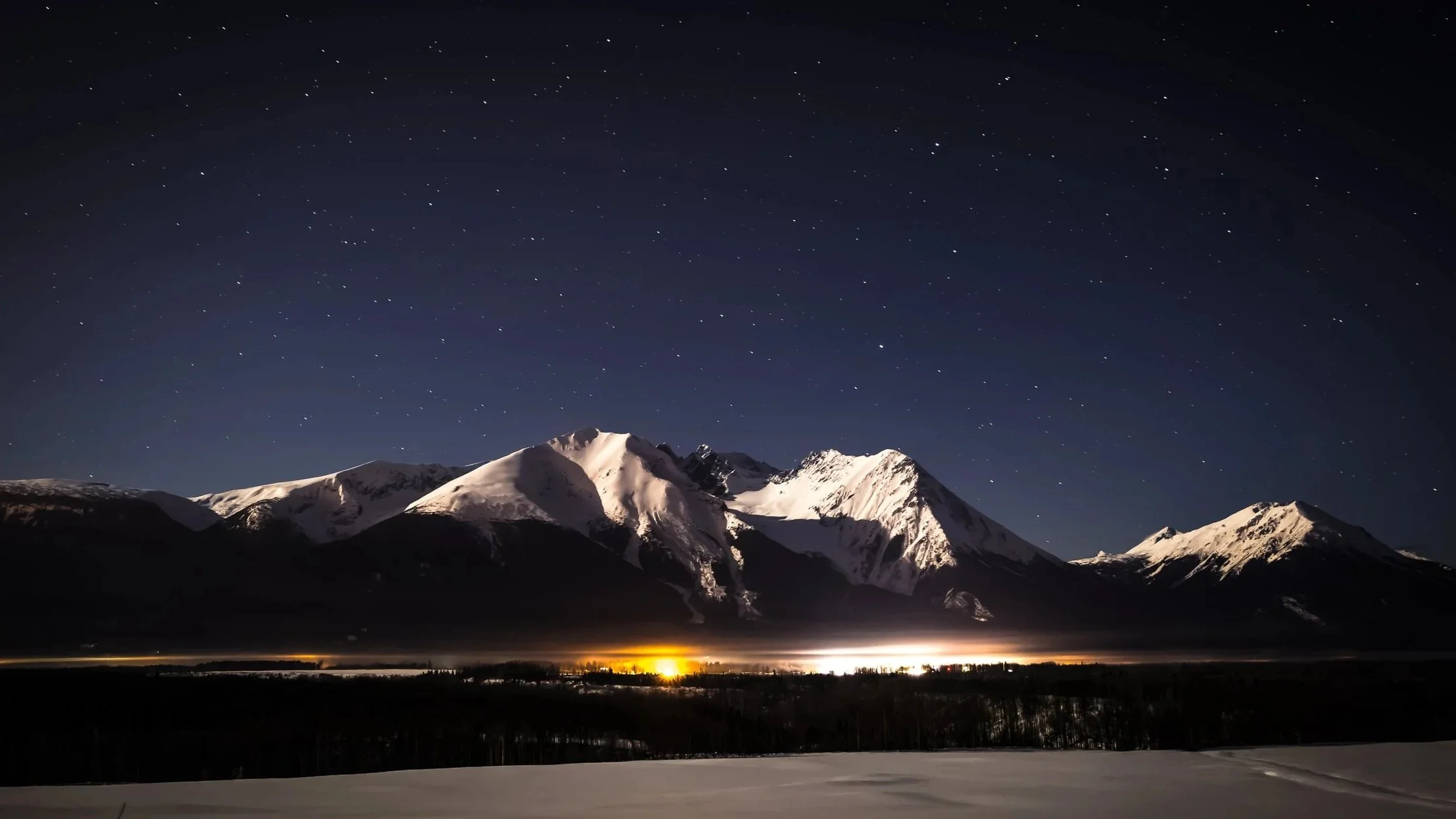 Snow-covered mountains at night with a starry sky and a glow of light at the horizon.