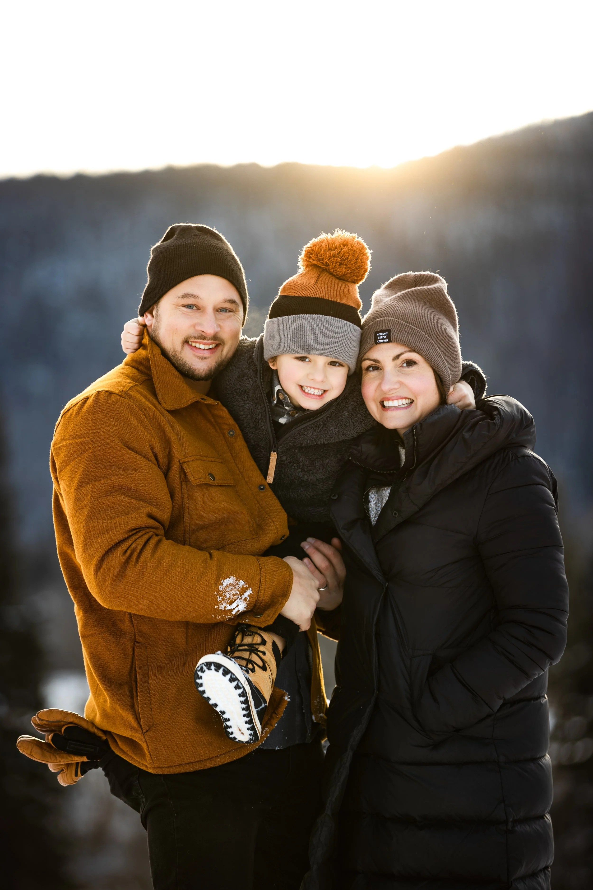 A happy family of three, dressed in winter clothing, outdoors during sunset, smiling and embracing each other, with snow on the ground and a mountain in the background.