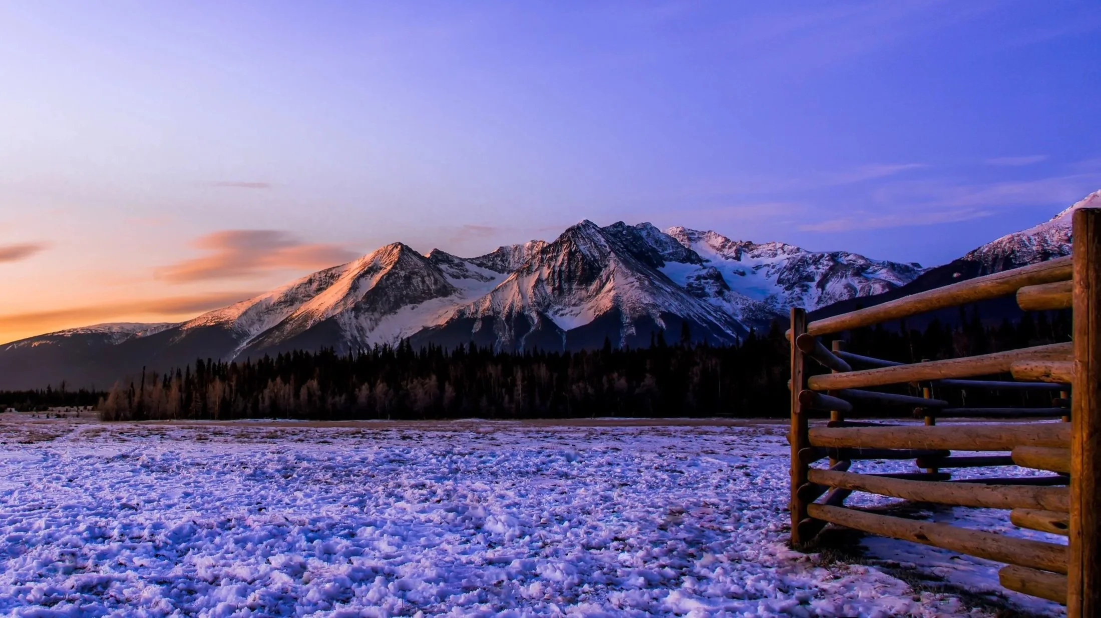 Snow-covered field with mountain range in the distance during sunset, and a wooden fence on the right side.