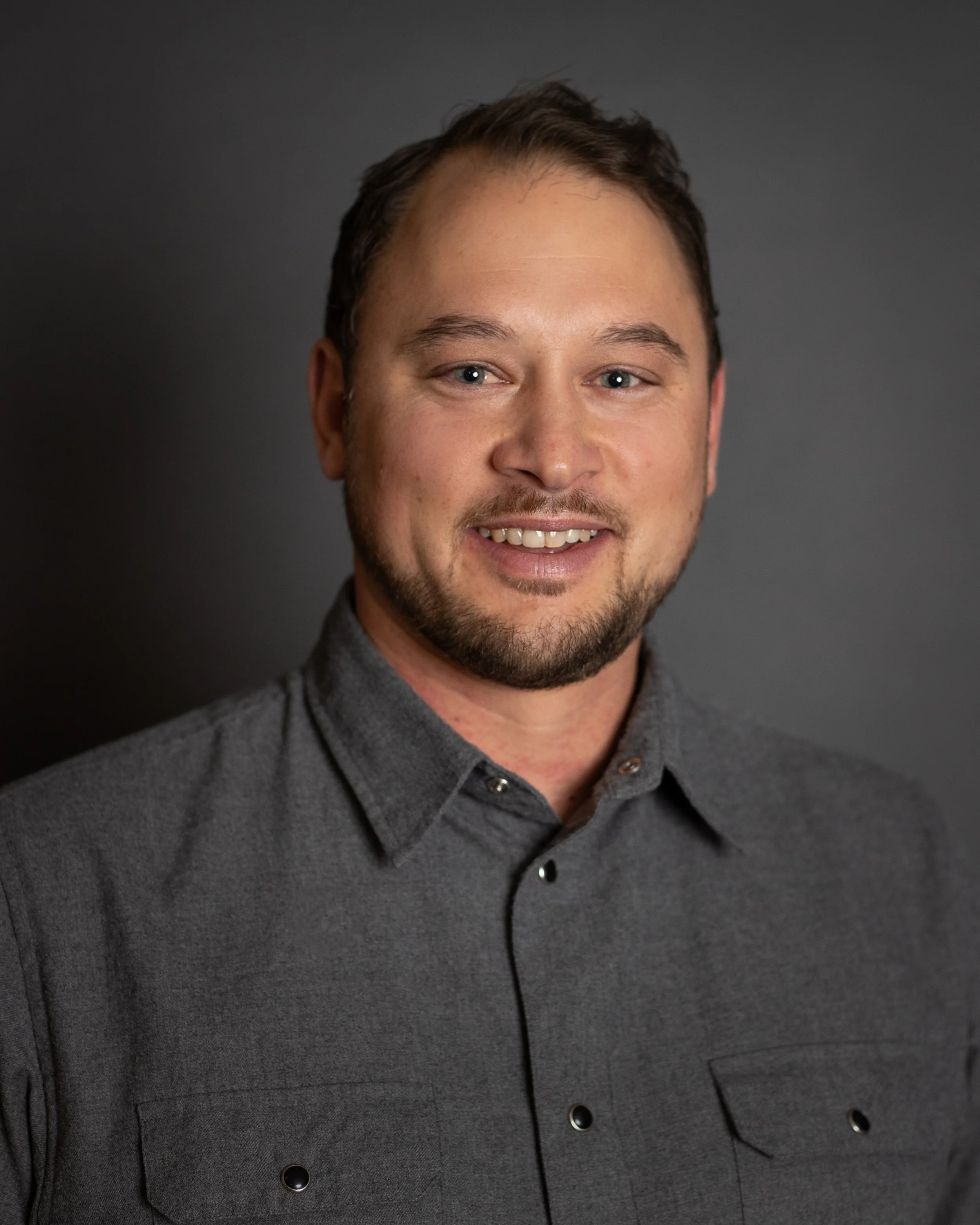 Portrait of a man with short dark hair, beard, wearing a gray button-up shirt, smiling against a dark background.