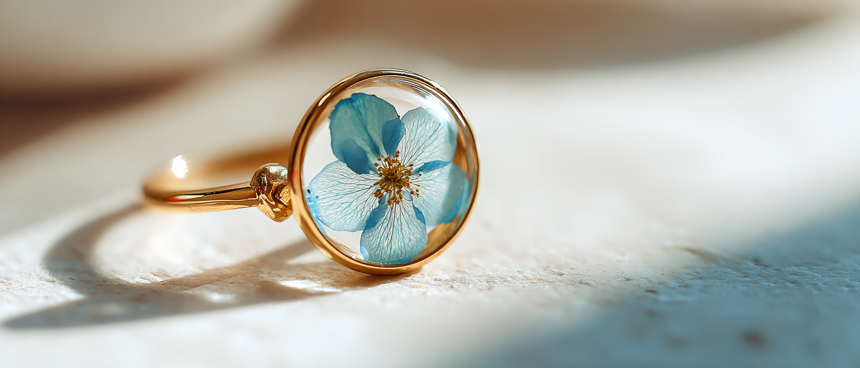 Custom flower jewelry in Washington D.C. A gold ring with a circular glass dome containing a blue flower, resting on a light surface with soft shadows.