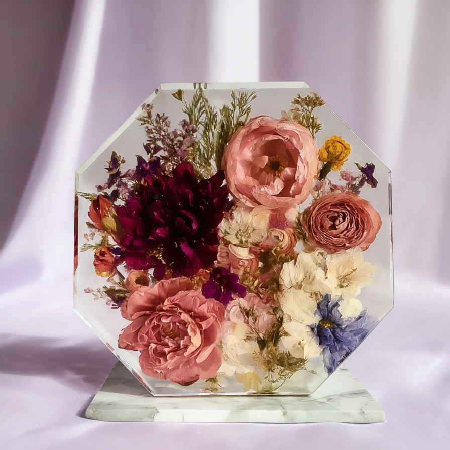 A clear octagonal glass sculpture filled with an arrangement of pink, purple, yellow, and white pressed flowers, mounted on a white marble base.