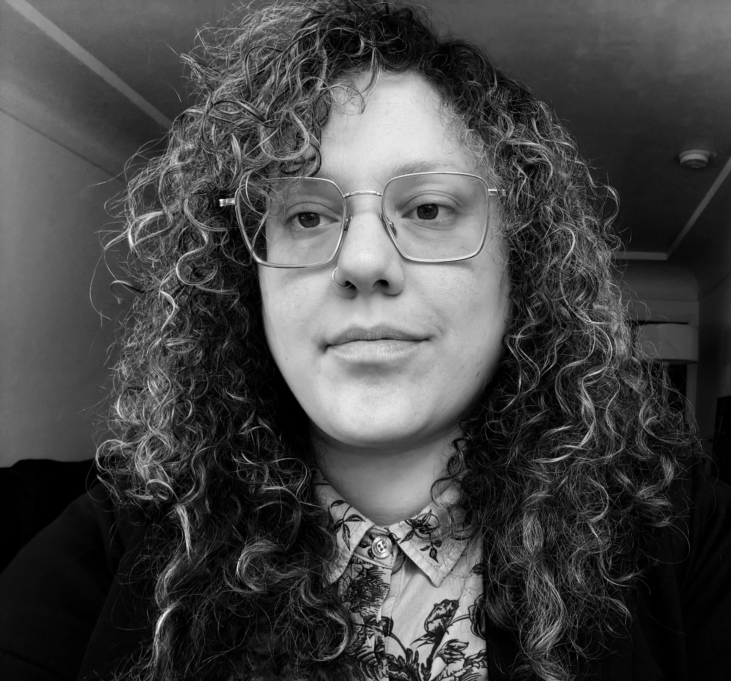 A woman with curly hair wearing glasses, a nose ring, a patterned shirt, and a jacket. She has a neutral expression and is indoors.