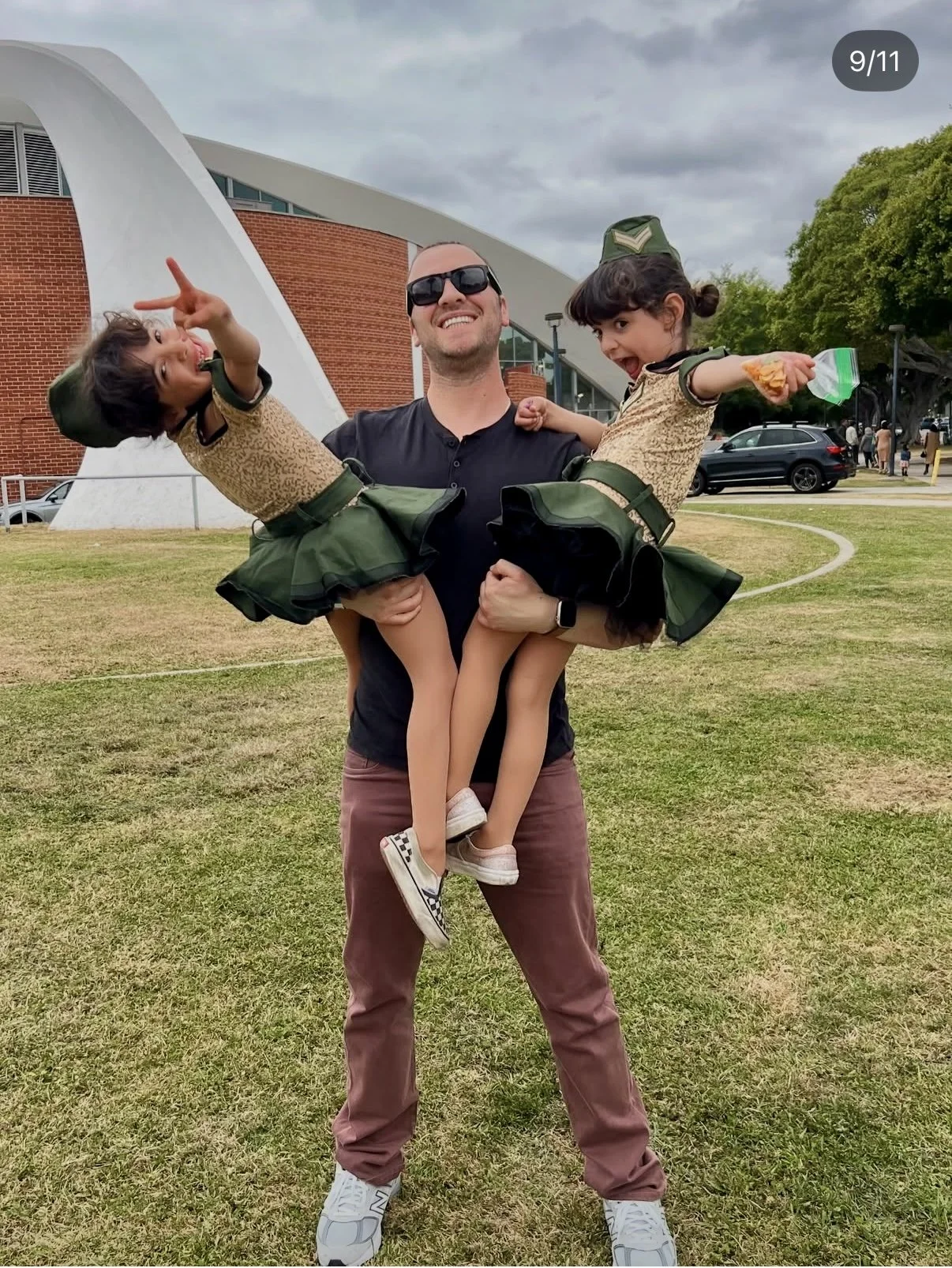 Tall white man with dark brown shaved hair with dark black sunglasses, black shirt and dark brown jeans is standing in front of a school holding 2, 7 year old girls with brown hair, gold and green dresses with green hats.