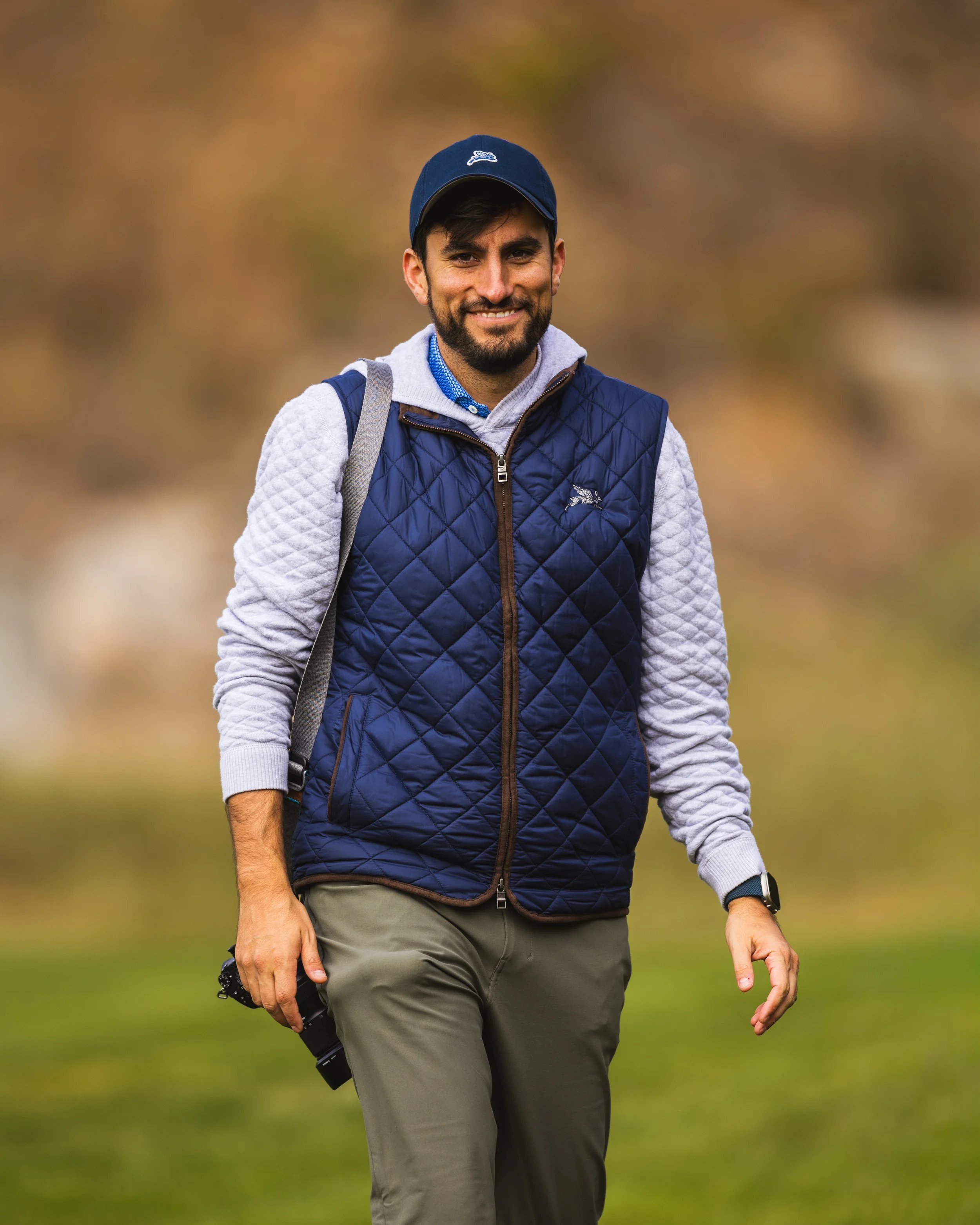 A smiling man with a beard walking outdoors on a trail, dressed in a blue quilted vest over a gray hoodie, wearing a blue cap and carrying a backpack.