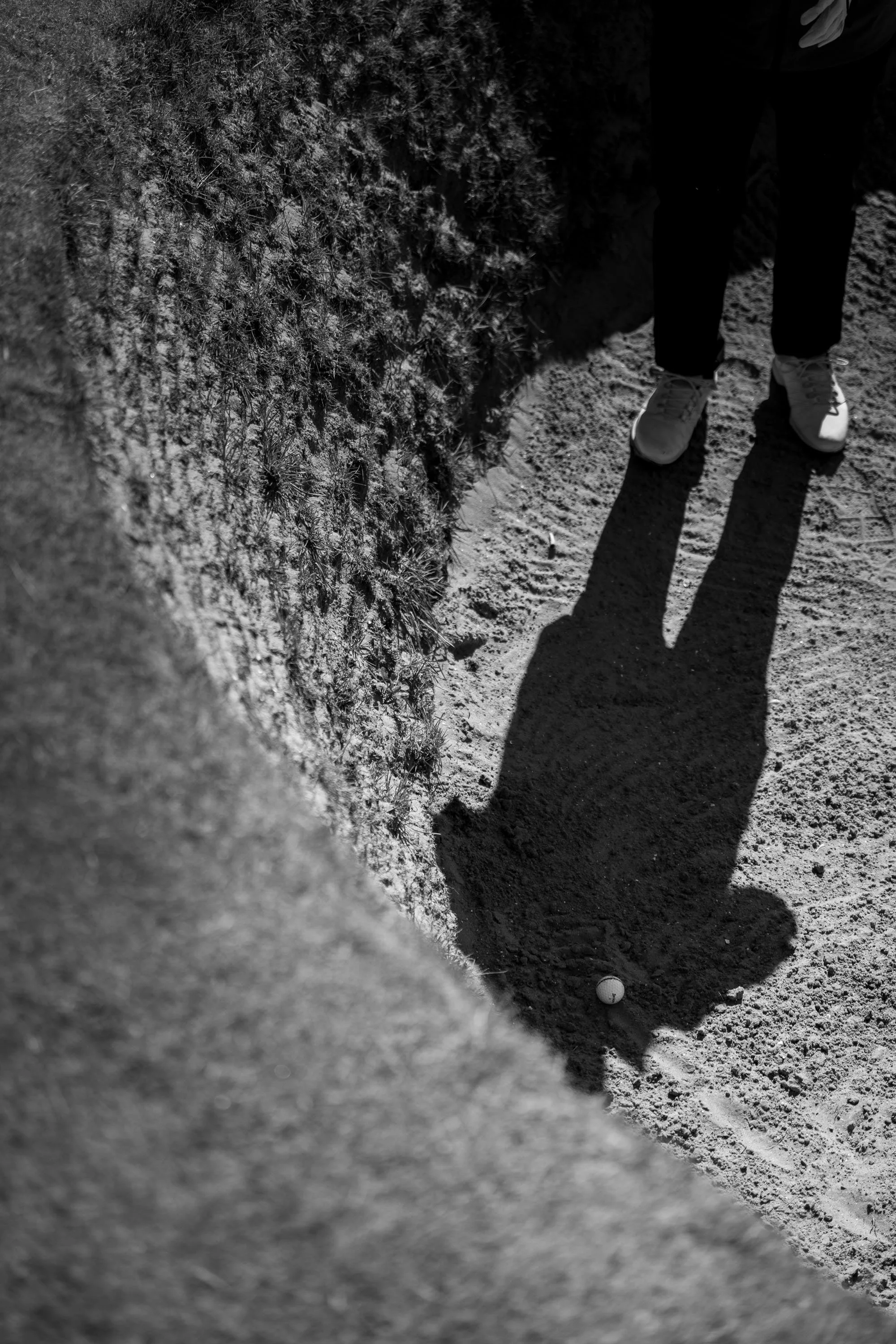 A black and white photo of a person standing in a bunker with their shadow cast long on the surface. The person's footwear is visible, and there is a small golf ball on the ground nearby. To the left, a hillside or mound covered in grass or plants.