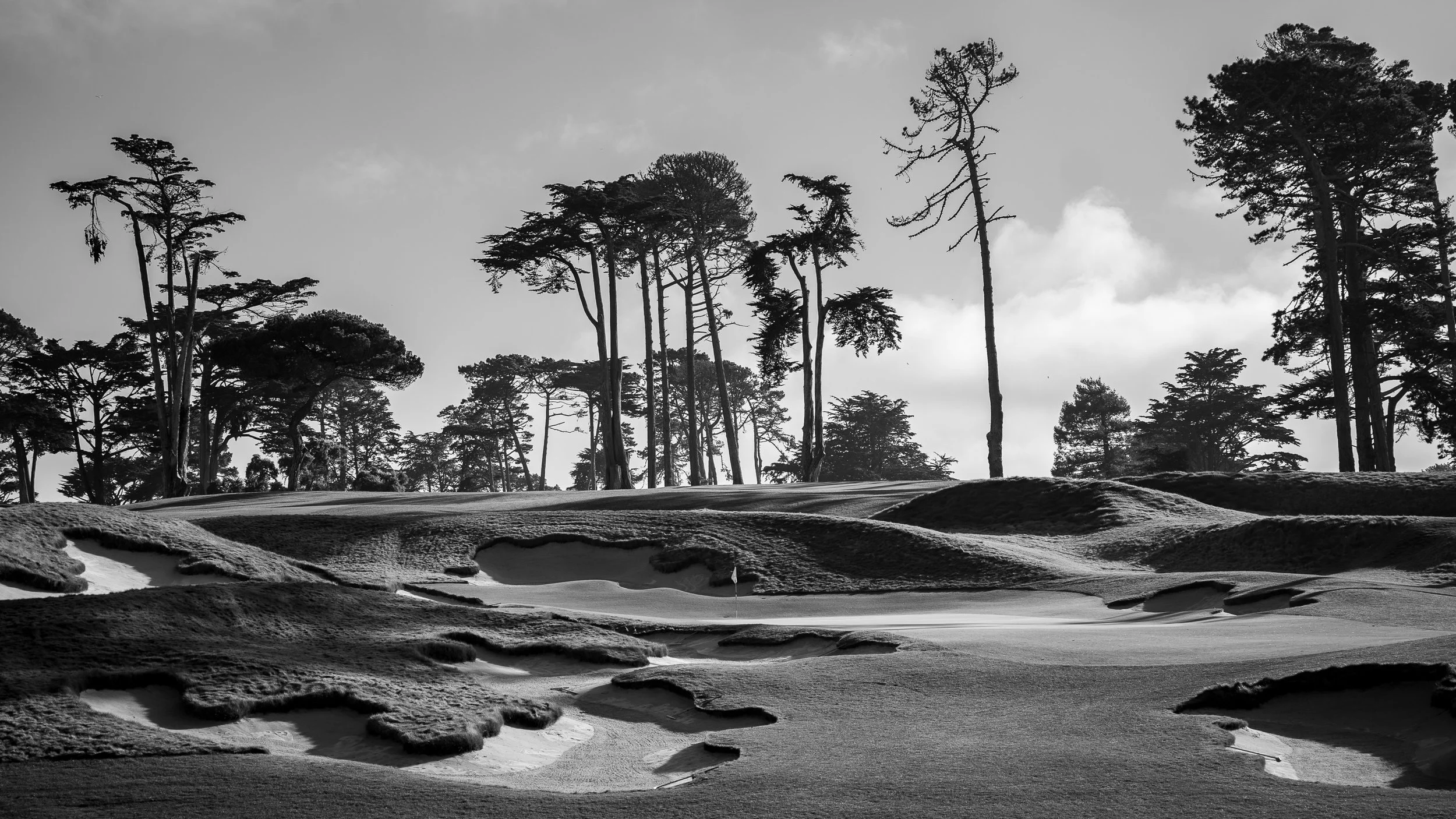 Black and white photograph of the 13th hole at Lake Merced with sand traps, rolling terrain, and tall, sparse trees in the background under a cloudy sky.