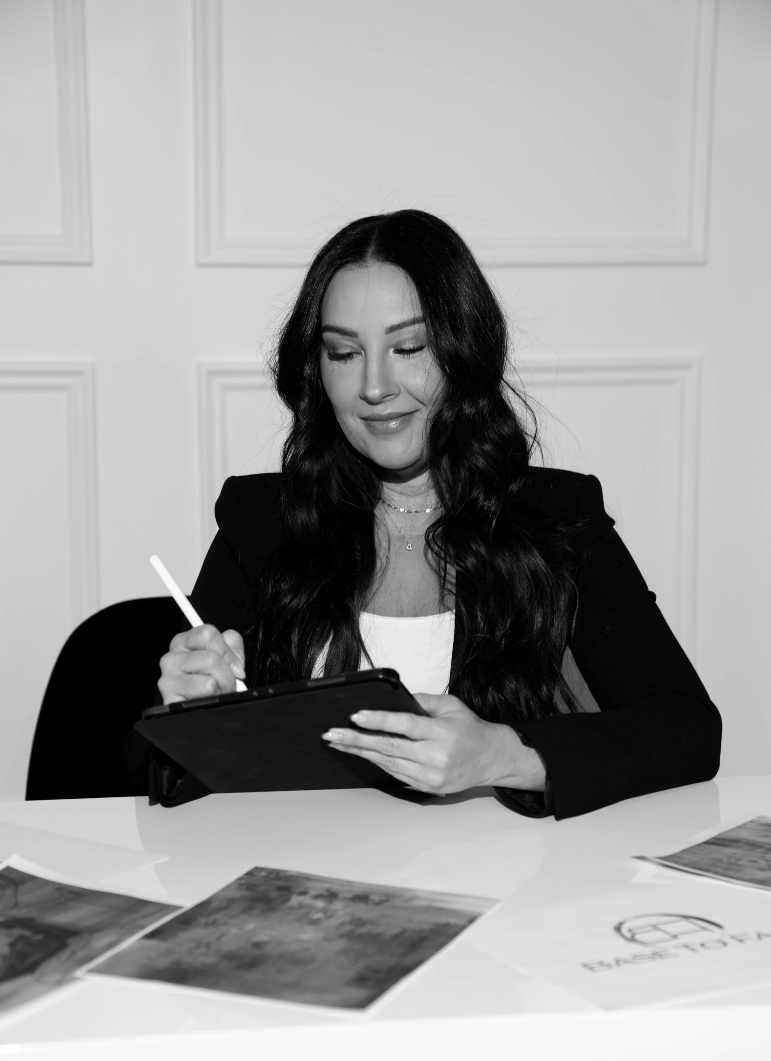 A woman with long dark hair, wearing a black blazer and white top, smiling and writing on a tablet with a stylus, seated at a table with papers and printed materials in front of her.