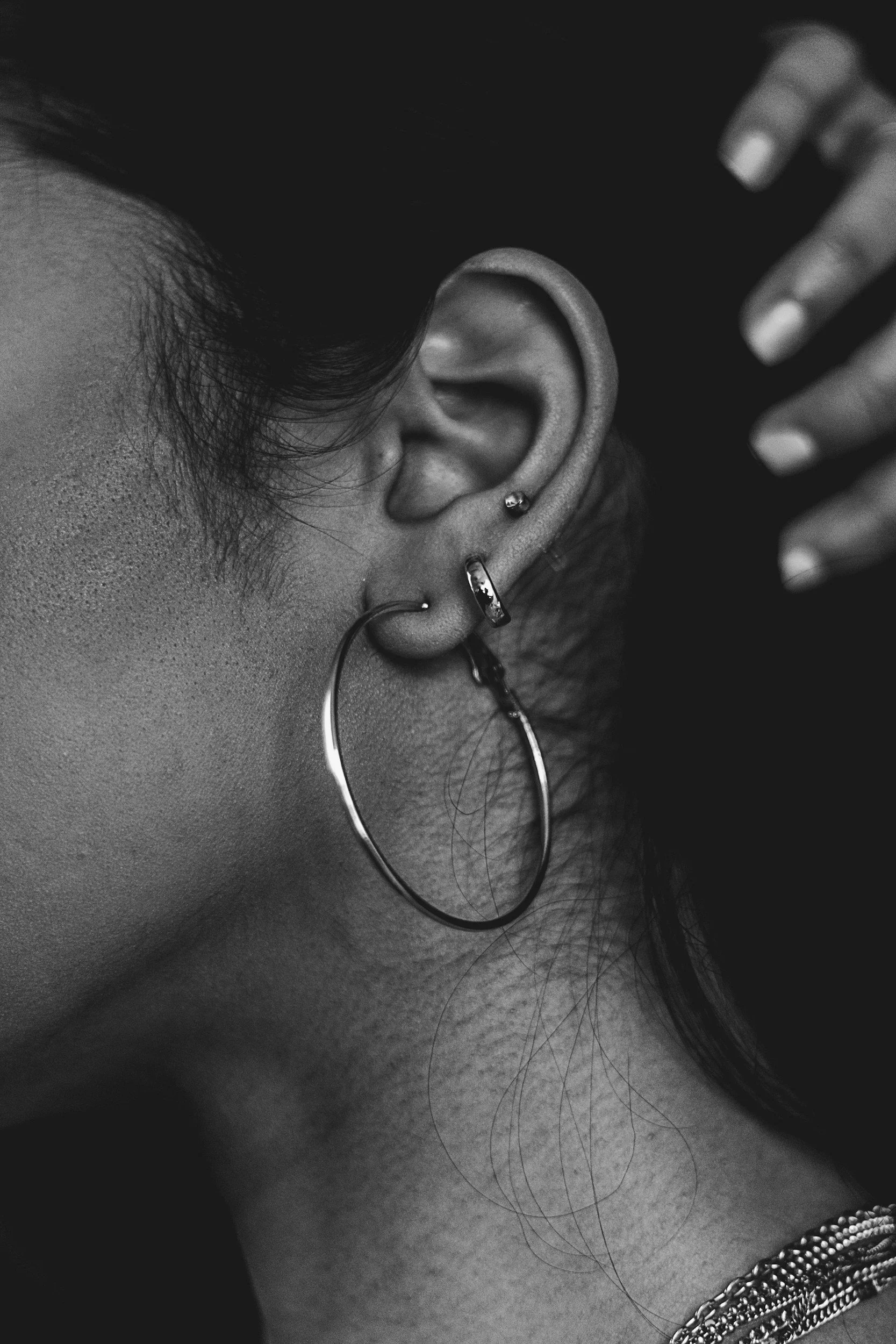 Close-up of a person's ear with multiple piercings, wearing hoop and stud earrings, with dark hair and a hand with manicured nails near the head, in black and white.
