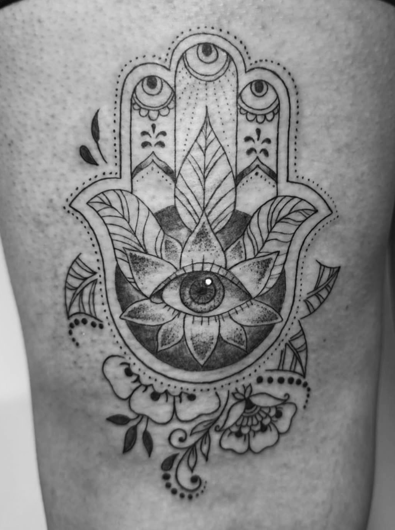 Tattoo of a Hamsa hand featuring multiple eyes, a central eye within a lotus flower, and floral elements.