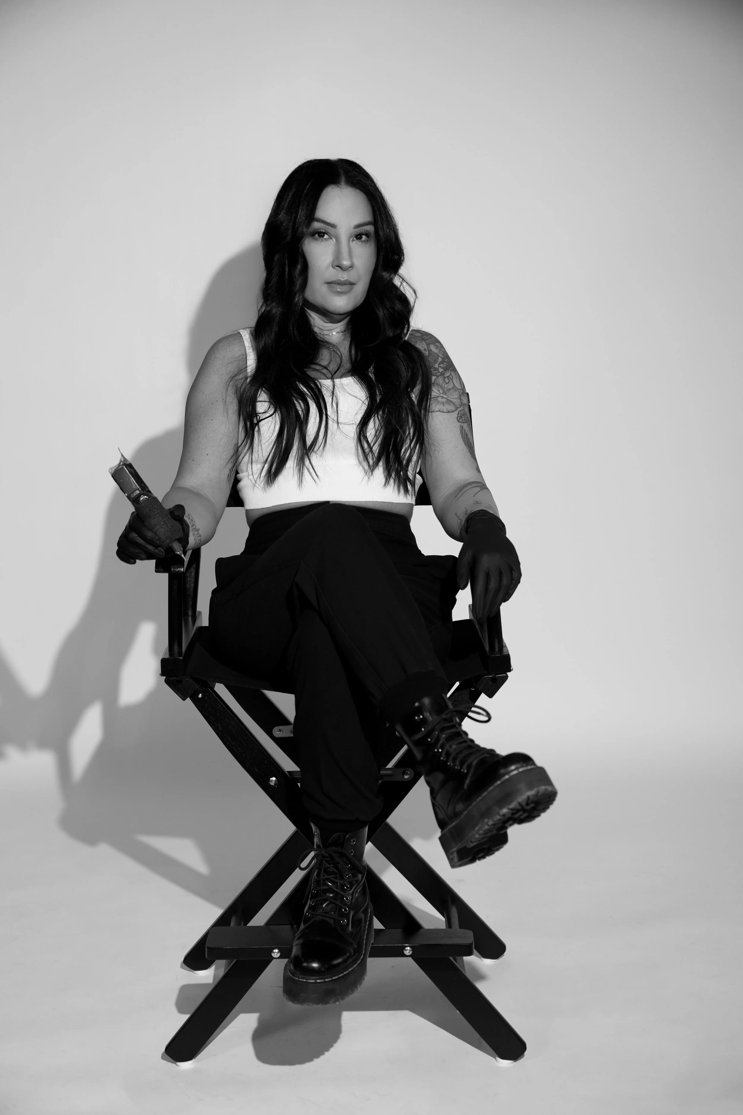A woman with long dark hair sitting on a director's chair, wearing a white sleeveless top, black pants, and black combat boots, holding a tattoo machine in her right hand in a studio setting with a plain background.