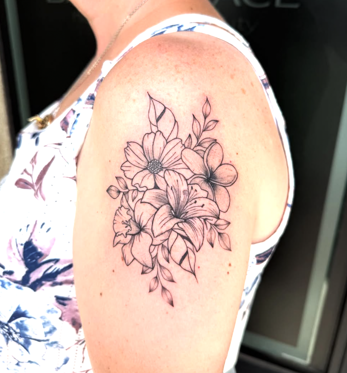 Black line floral tattoo featuring lilies, a daisy, and leaves on a person's upper arm.