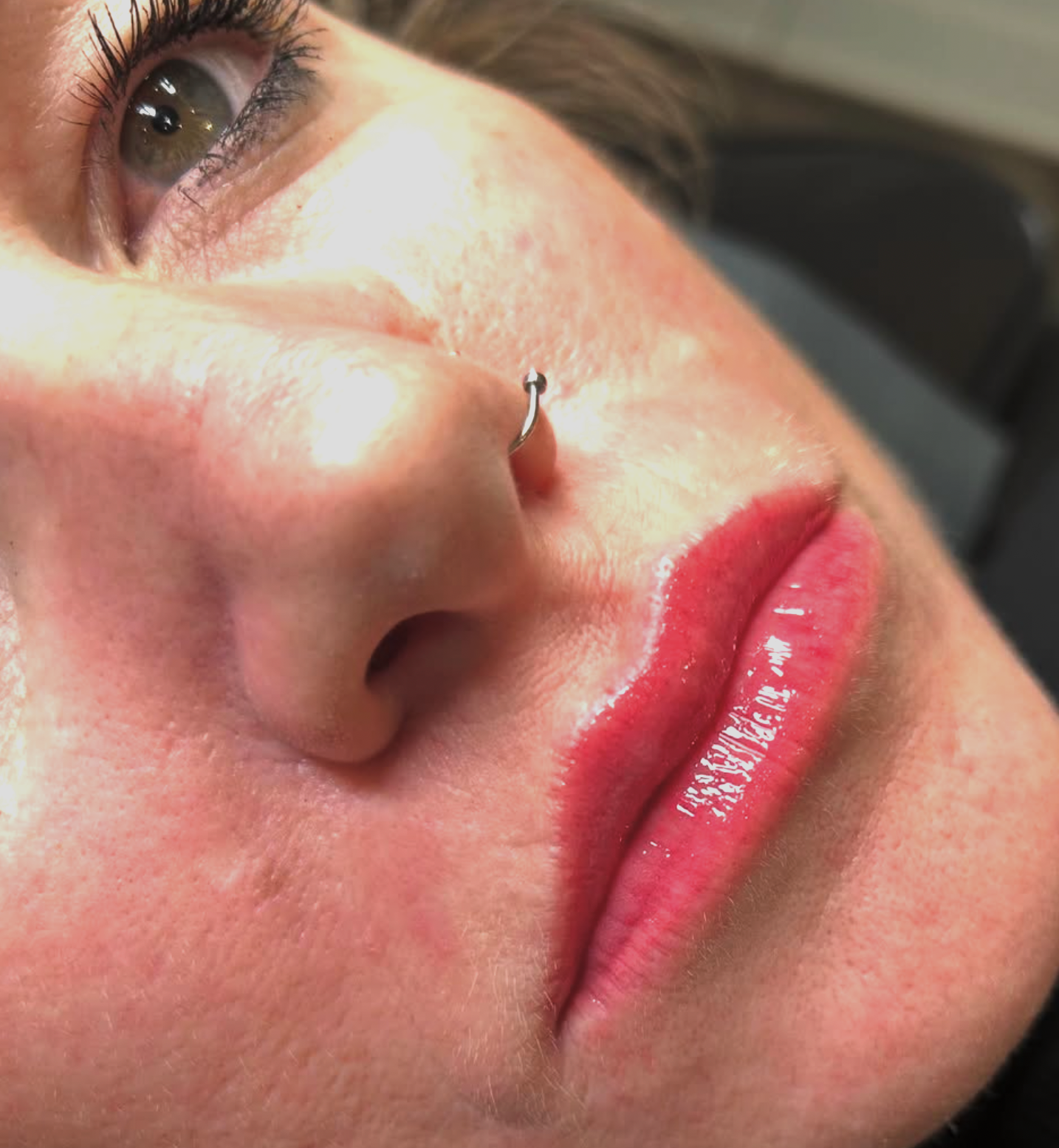Close-up of a woman's face showing her eye, nose with a septum piercing, and lips with shiny lipstick.