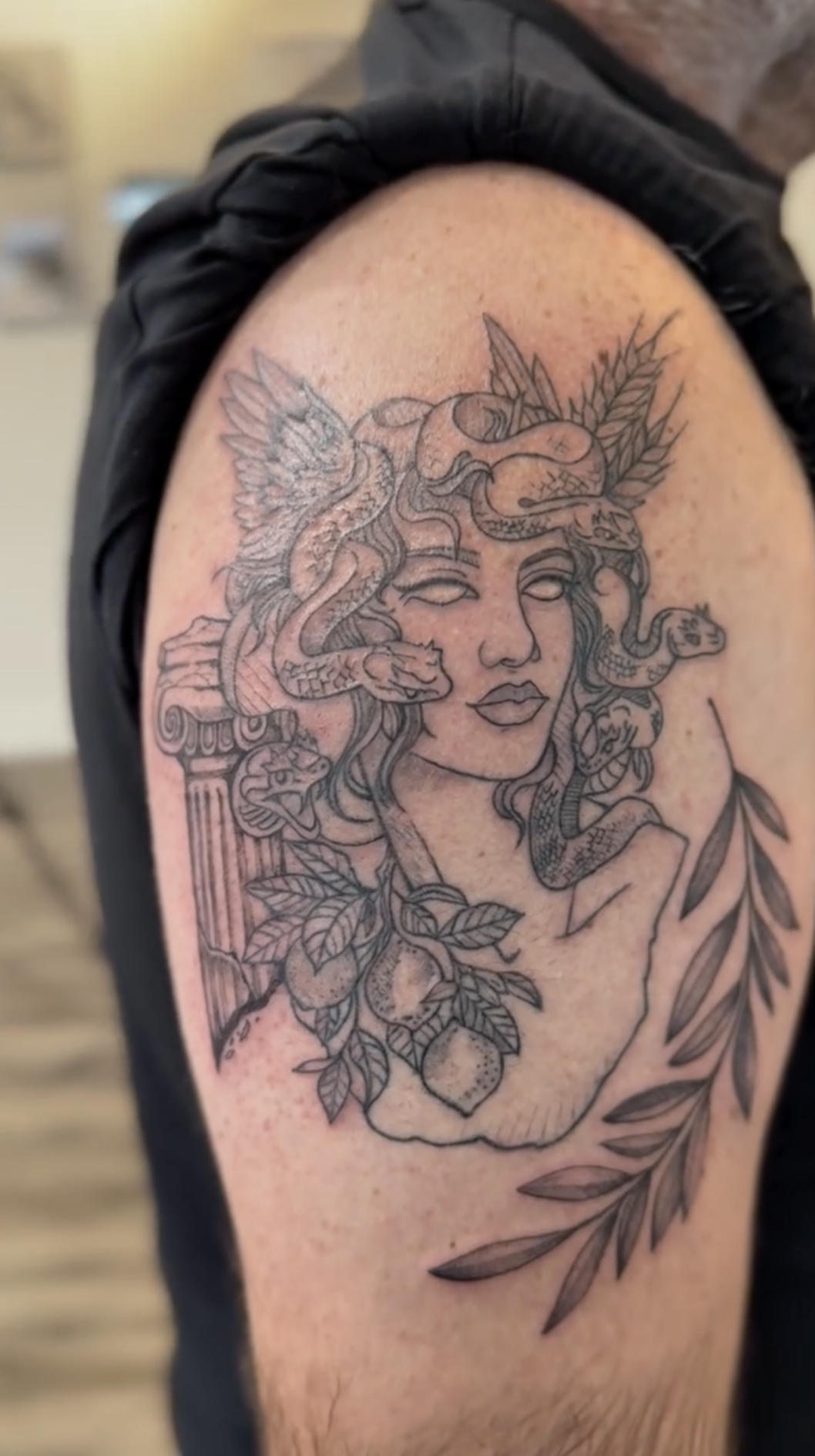 Line art tattoo of a woman’s face with snakes, a bird, and foliage, including lemons and leaves, on upper arm.