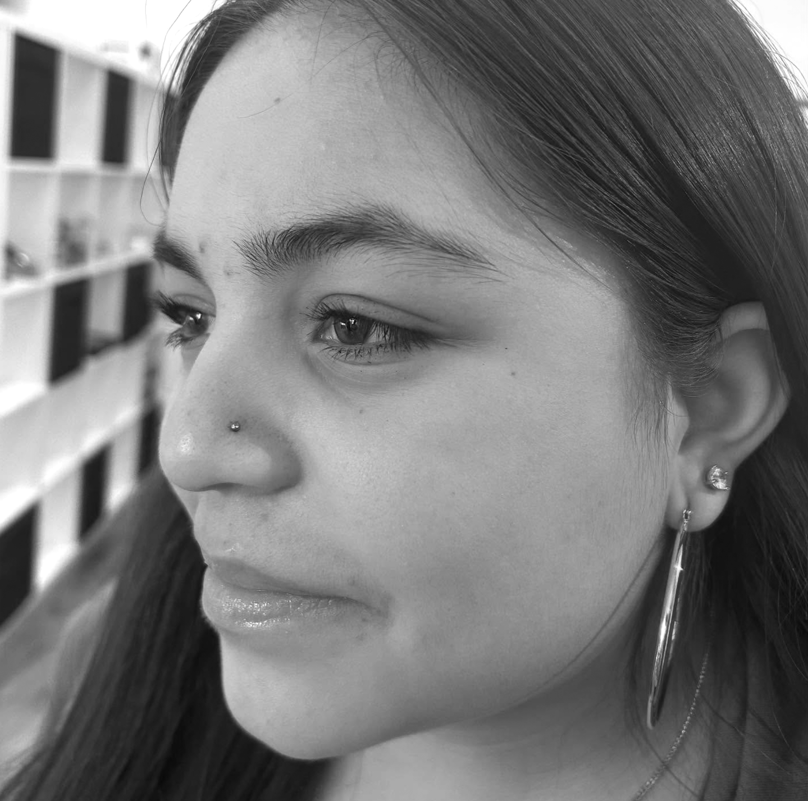 Black and white close-up of a young woman's face, showing her left profile with straight dark hair, pierced nose, and earrings. She is looking to the left with a neutral expression.