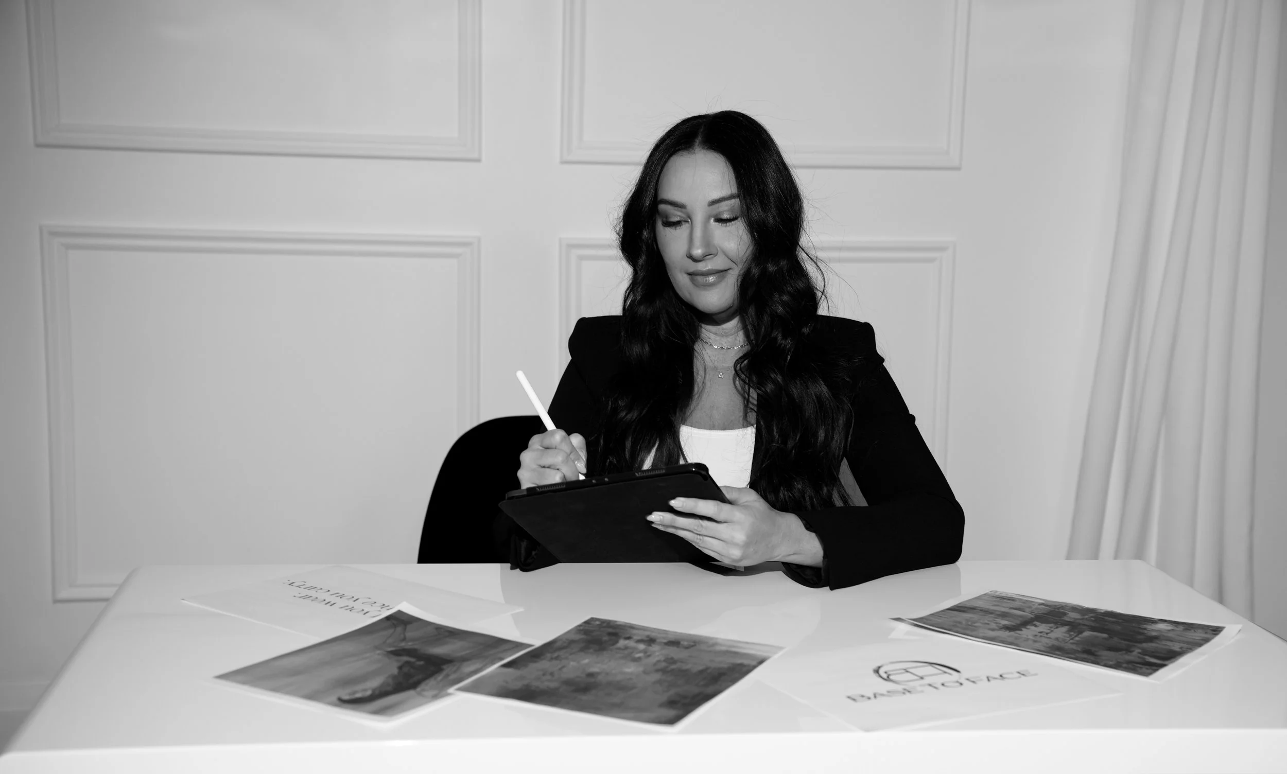 A woman with long dark hair in a black blazer and white top sits at a white table, writing on a digital tablet with a stylus. There are photographs and documents spread out on the table in front of her.