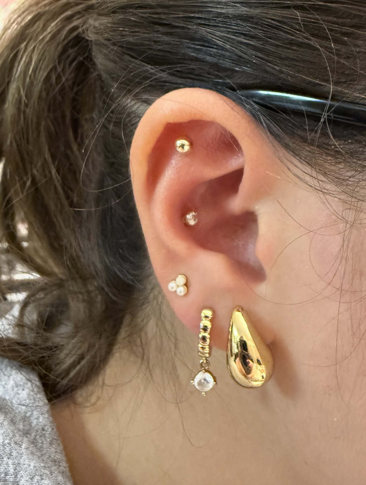 Close-up of a person's ear with multiple earrings including small studs, a hoop with a dangling gem, and a large gold teardrop-shaped earring.