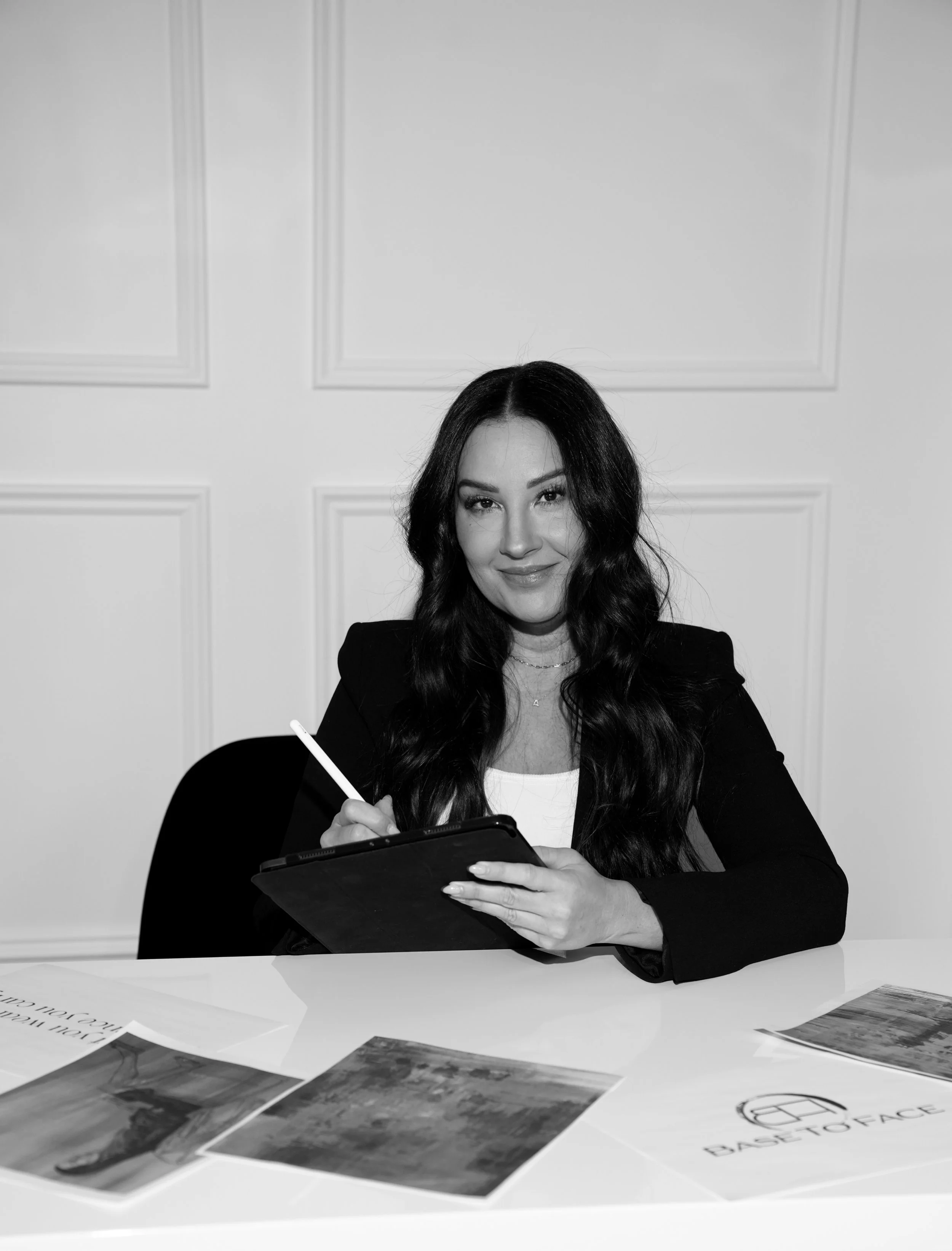 A woman with long dark wavy hair, smiling, sitting at a table with photographs and documents, holding a tablet and stylus.