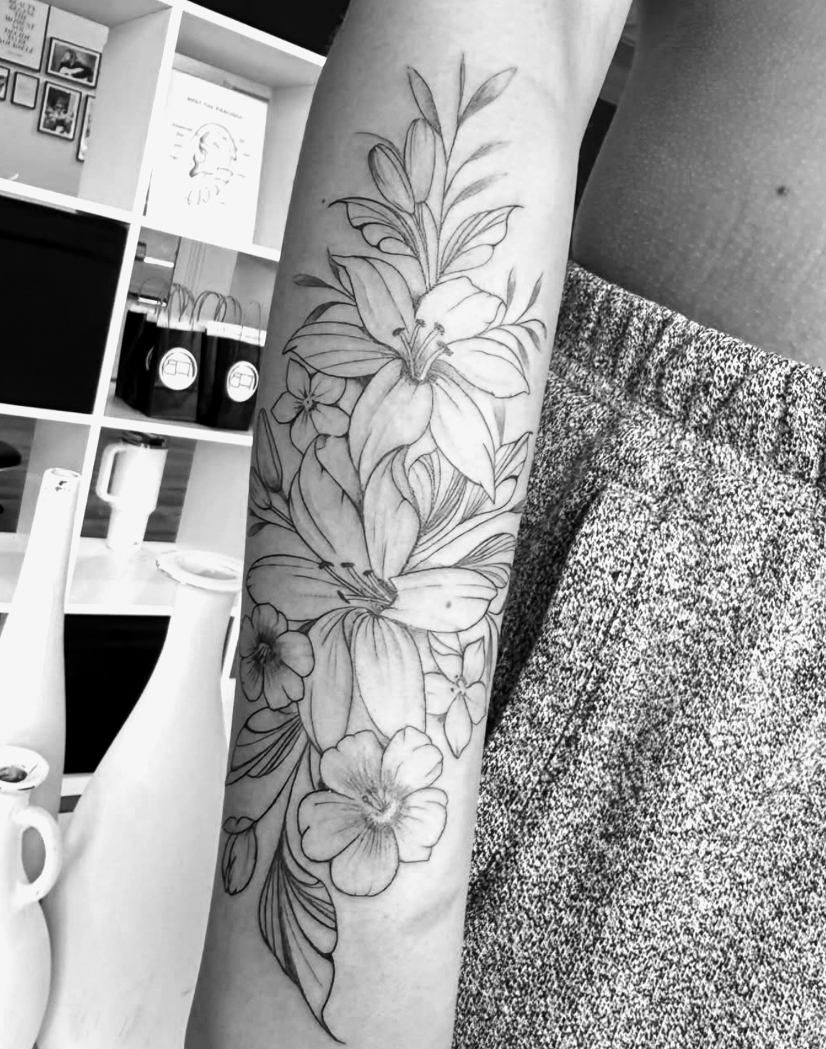 A black and white photo of a tattoo on a person's forearm featuring multiple lilies and other flowers.