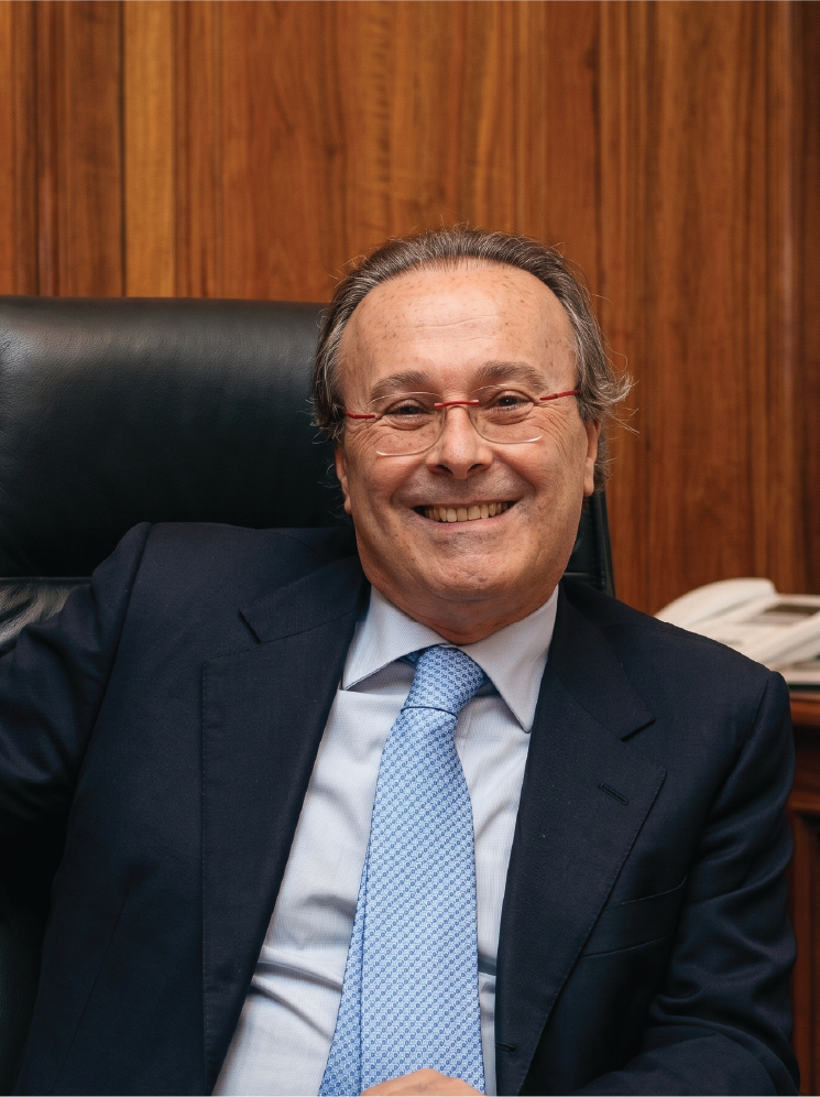 Dr. Piergiorgio Spaggiari, a smiling middle-aged man wearing glasses, a dark suit, light blue tie, sitting in an office chair against a wood-panelled wall, with a telephone on the desk behind him.