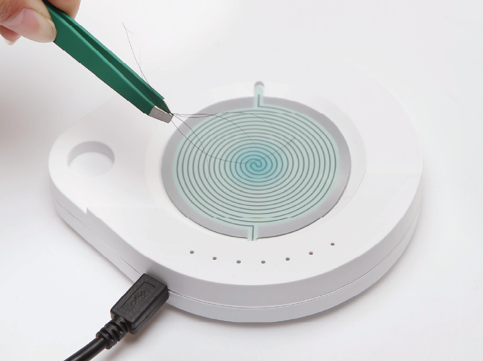 A hand holding a green pen drawing spiral lines on a modern, circular, white device with a spiral pattern in the center, connected by a black USB cable.