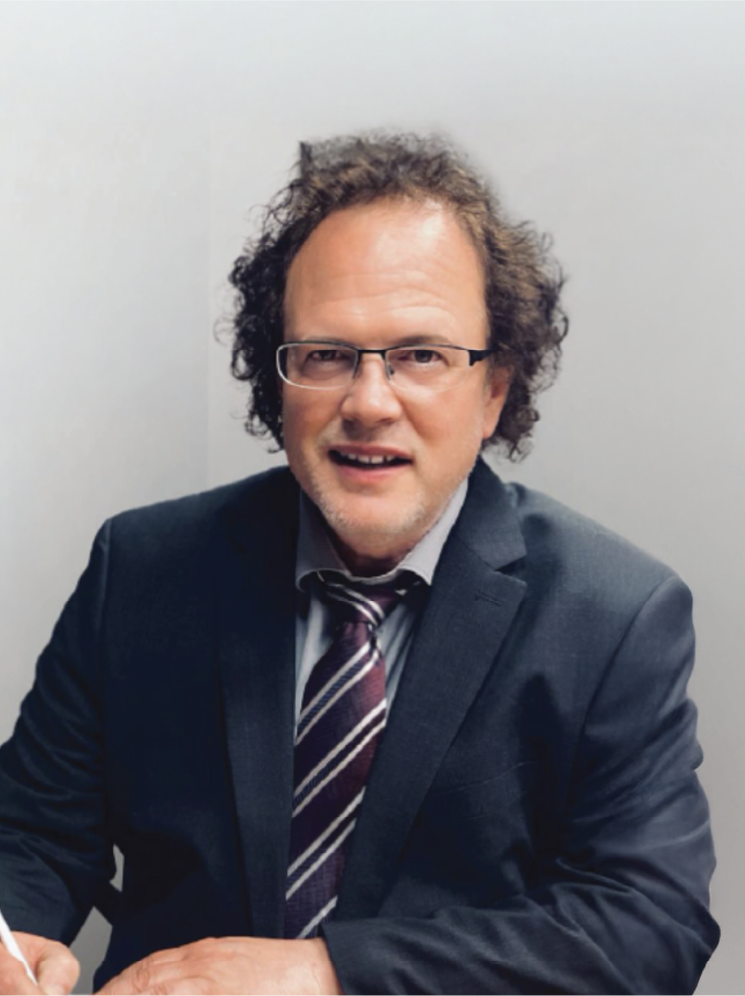 Dr Marcus Stanton, a middle-aged man with curly hair, glasses, and a beard wearing a dark suit, dress shirt, and striped tie, is sitting at a table in front of a plain white wall.