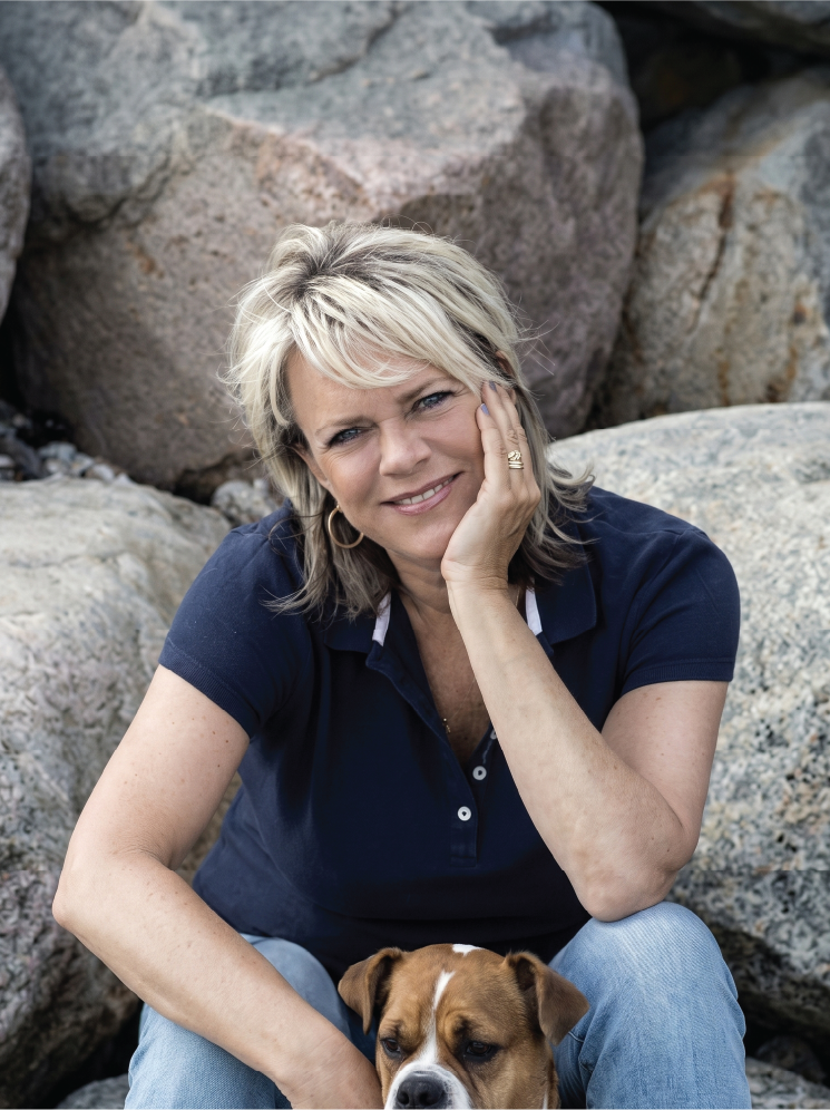 Lene Hansson, a woman with blonde hair sitting outdoors next to large rocks, smiling, with a brown and white dog.