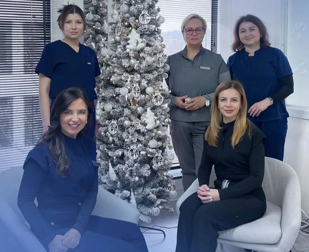 Five women, three in medical scrubs, posing around a decorated white Christmas tree in an office.