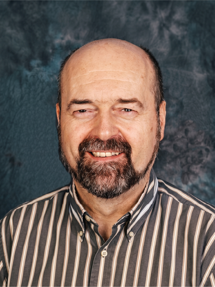 Dr Folker Meissner, a middle-aged man with a beard and moustache, smiling slightly, wearing a striped button-up shirt, against a textured blue background.