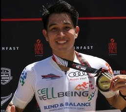 Smiling young male triathlete with short dark hair, wearing a white cycling jersey with Cell Wellbeing logos, holding a medal, standing in front of a black backdrop with red and white lettering.