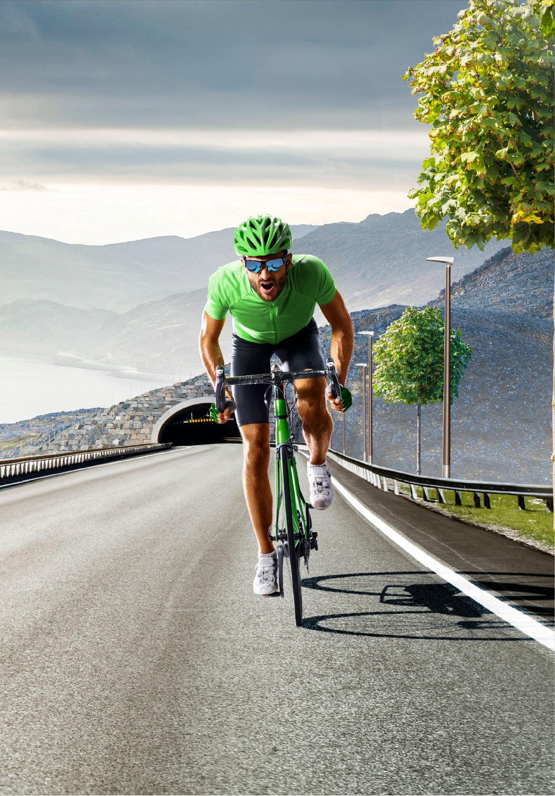 A man in a green shirt and helmet riding a bicycle on a mountain road with trees and a tunnel in the background.