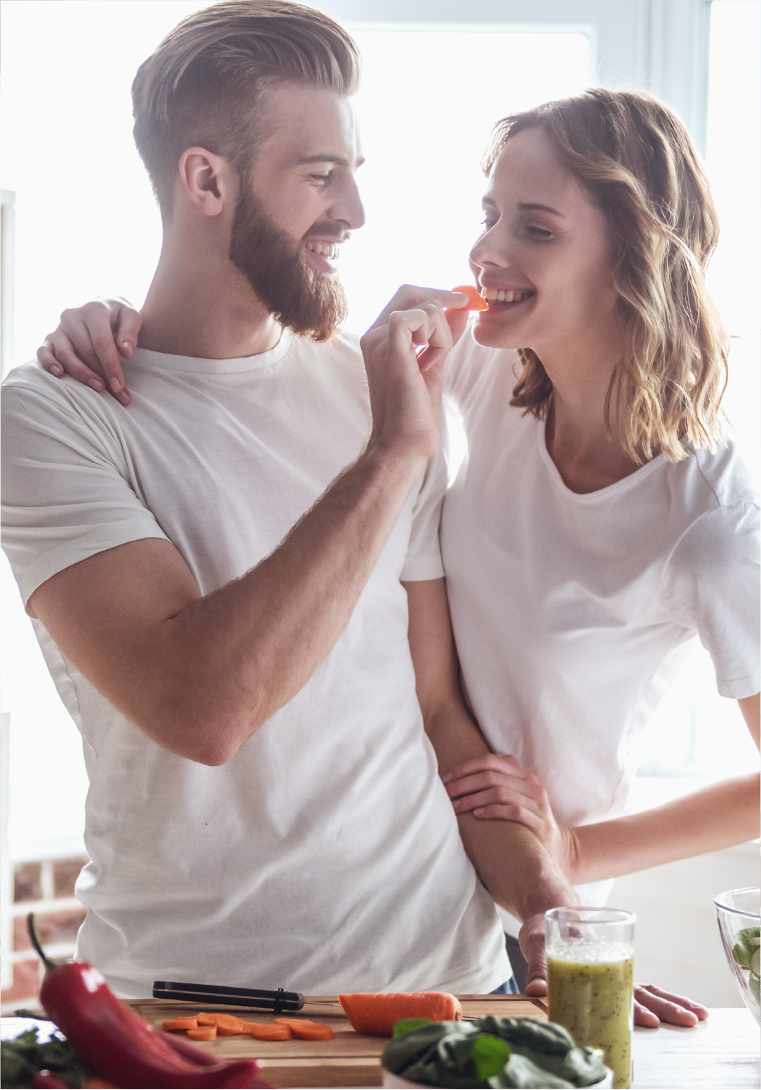 A man feeding a woman a piece of vegetable in a kitchen with fresh vegetables and a smoothie on the counter.