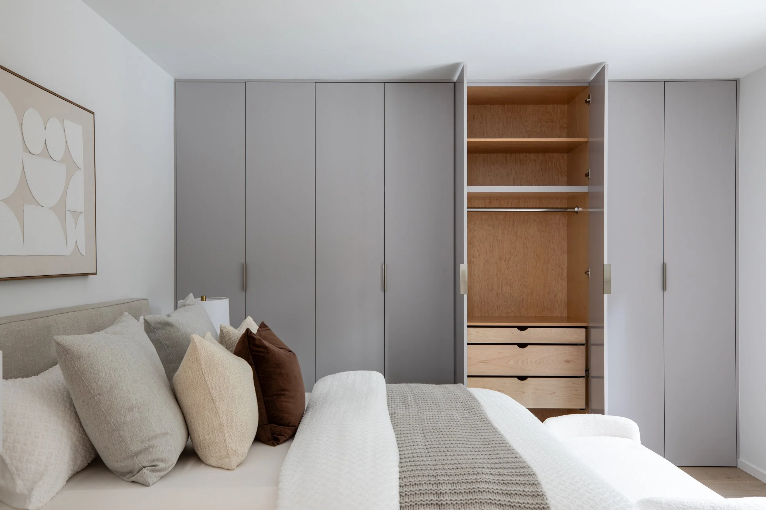 Modern bedroom with a bed covered in neutral-colored pillows and a white blanket, next to a built-in closet with gray doors and open drawers.