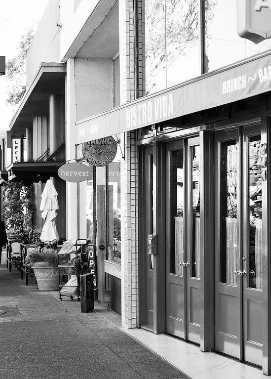 Black and white photo of a sidewalk outside a restaurant named Bistro Vida, with outdoor seating and umbrellas, next to a shop named Harvest.