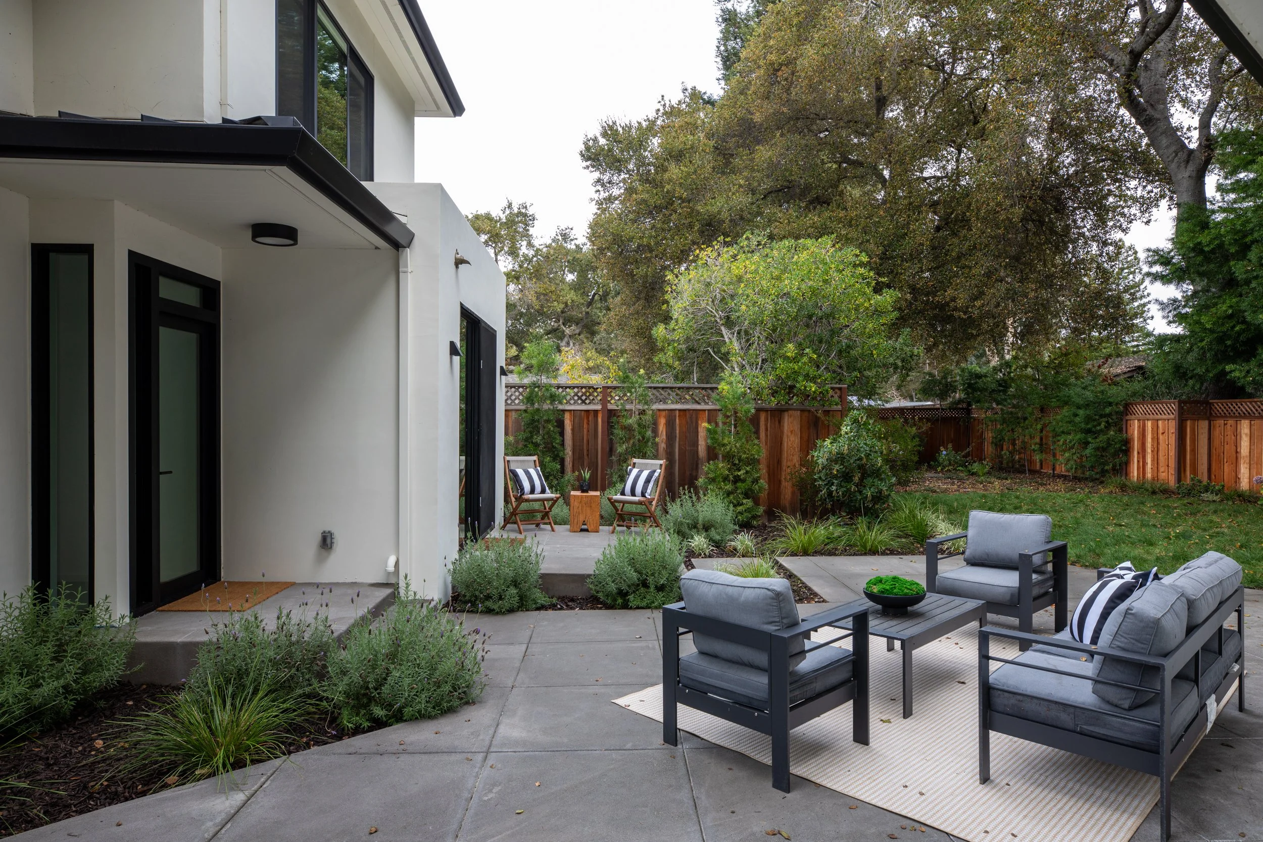 Modern backyard patio with outdoor furniture including a gray cushioned sofa, chairs, and a black coffee table on a striped rug. There are two wooden chairs with striped pillows on a slightly elevated deck, surrounded by lush greenery and a wooden fe