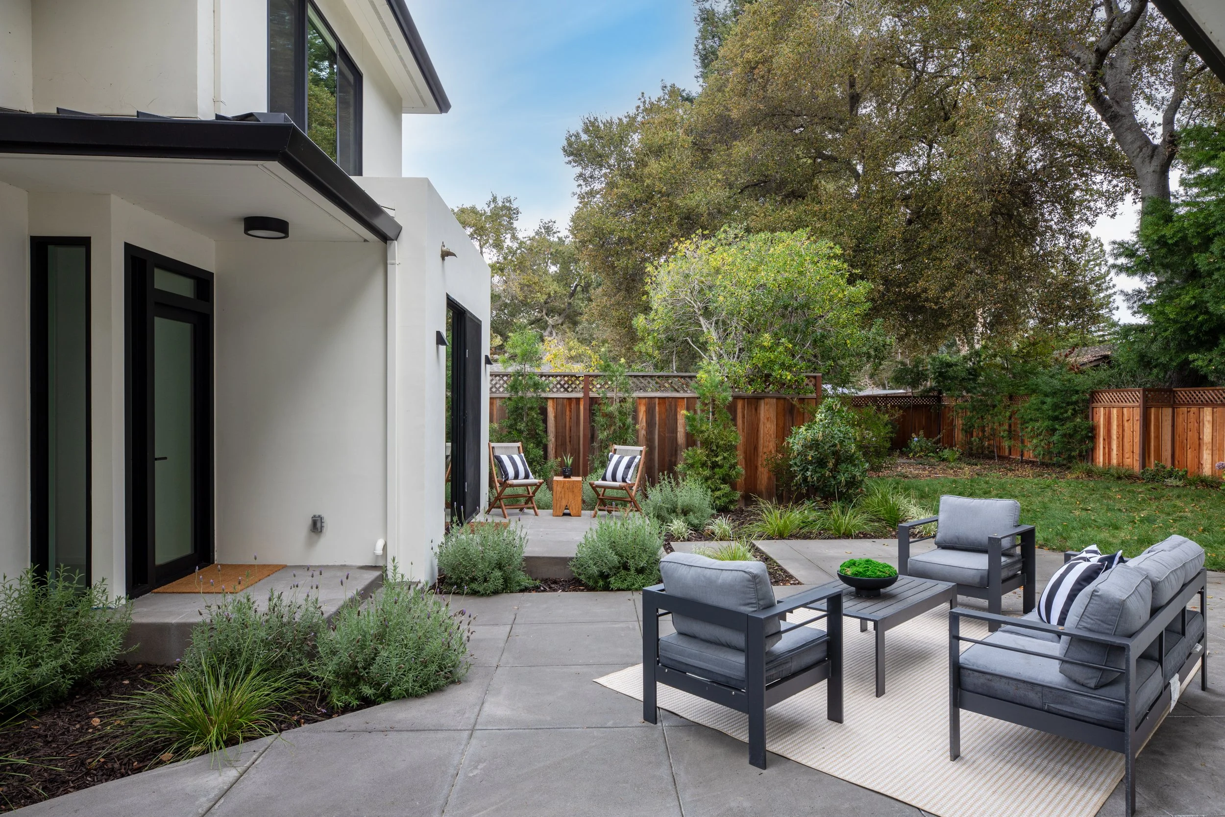 Modern backyard patio with outdoor seating, including a gray sofa and armchairs, a rug, and a garden with trees and plants, enclosed by a wooden fence.