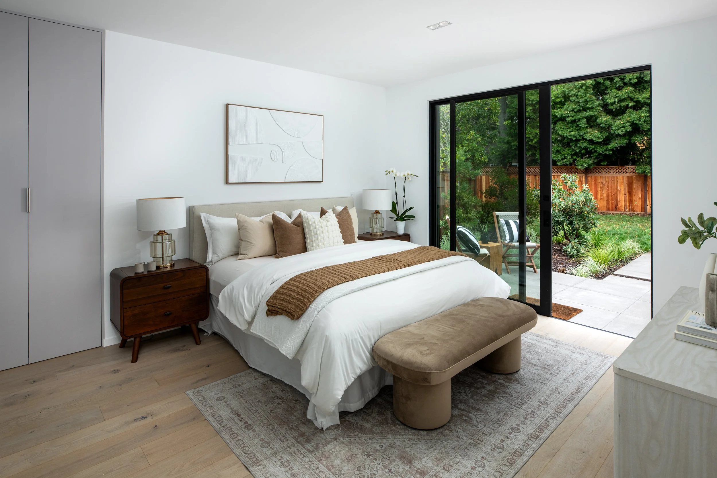 Bedroom with a large bed, nightstands, lamps, a bench at the foot, and sliding glass doors leading outside to a garden.