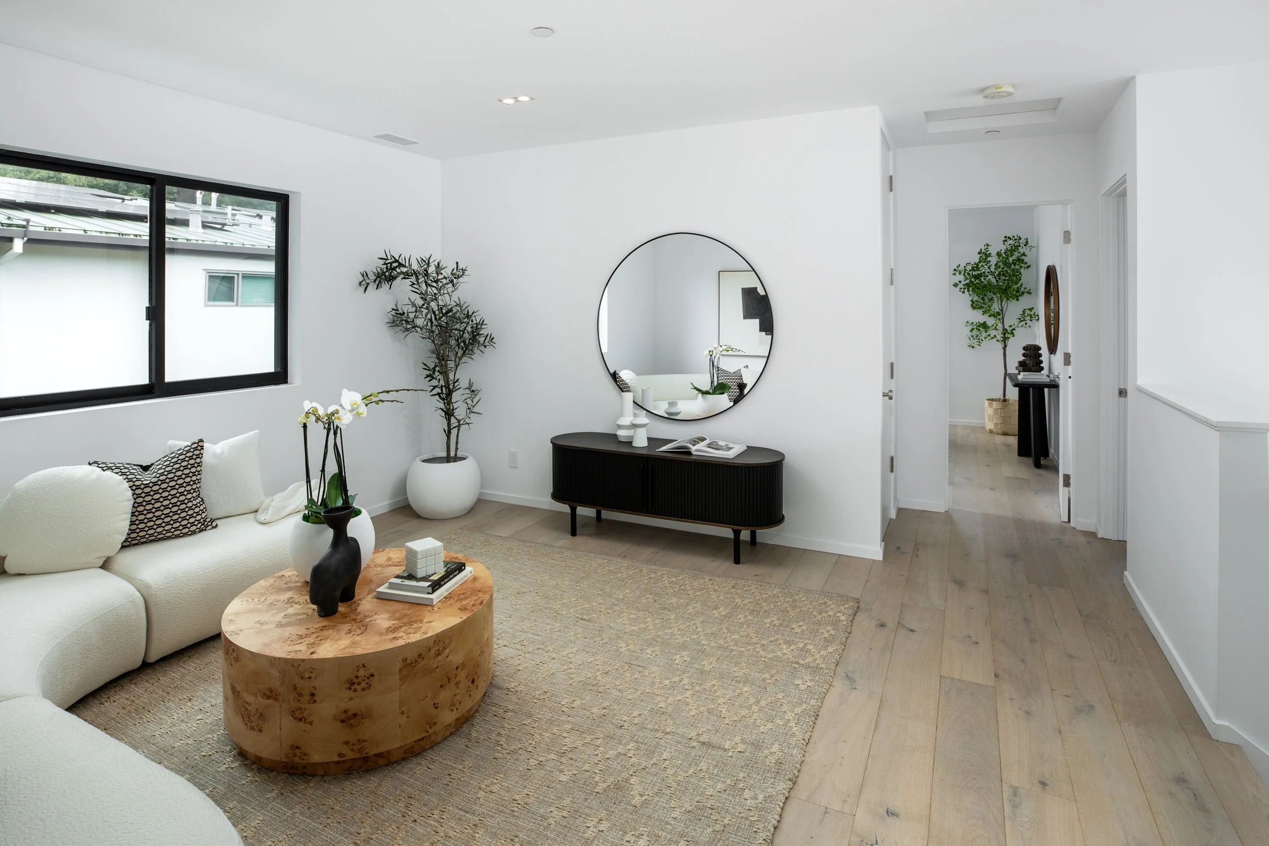 Minimalist living room with white sofa, wooden coffee table, black-framed window, large potted plant, round wall mirror, black sideboard with decorative items, light hardwood floors, white walls, and a hallway with another potted plant.
