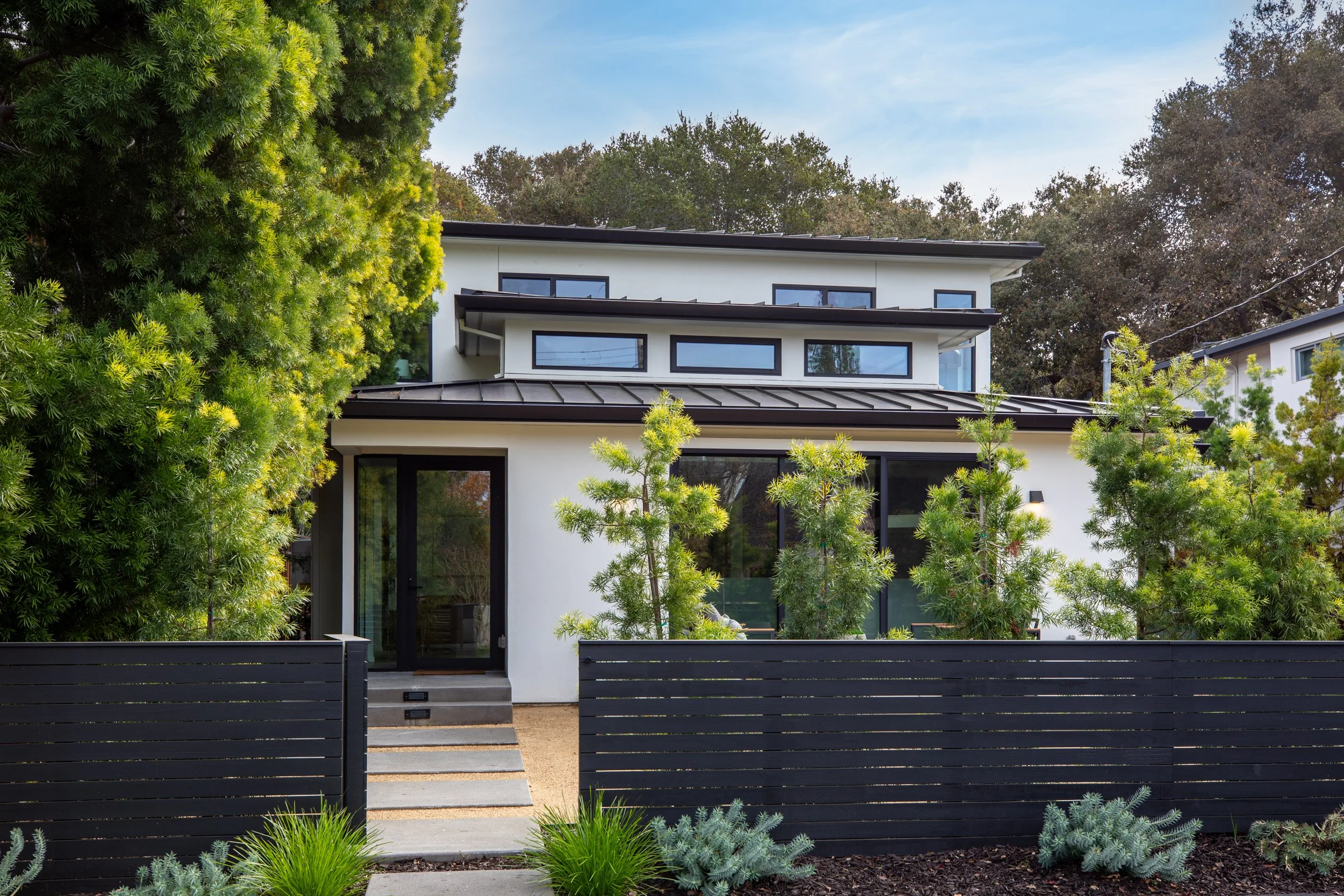 Modern white multi-story house with black accents, large windows, and a black wooden fence at the front, surrounded by green trees and plants.