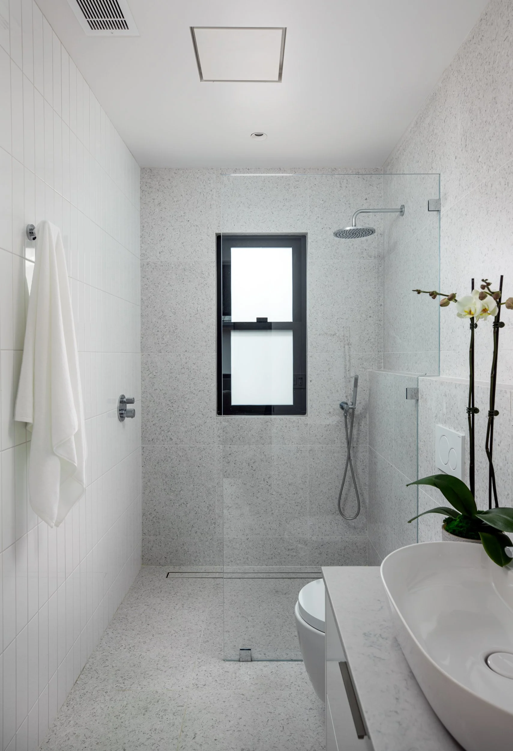 Modern minimalist bathroom with walk-in shower, frosted window, white sink with orchid plant, and white tiled walls.