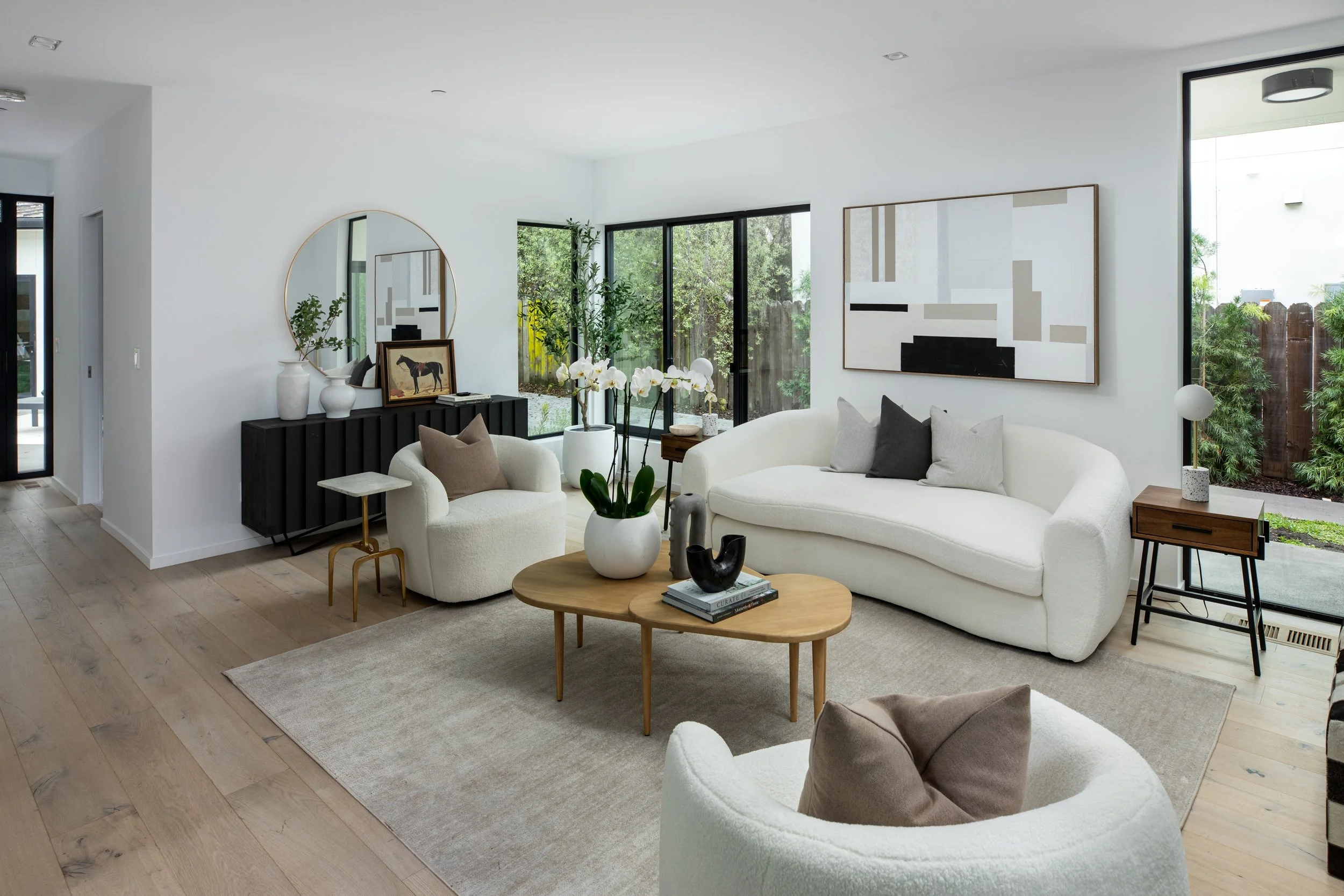 Modern living room with white couches, wooden tables, potted plants, and abstract wall art, bright natural light from large windows.